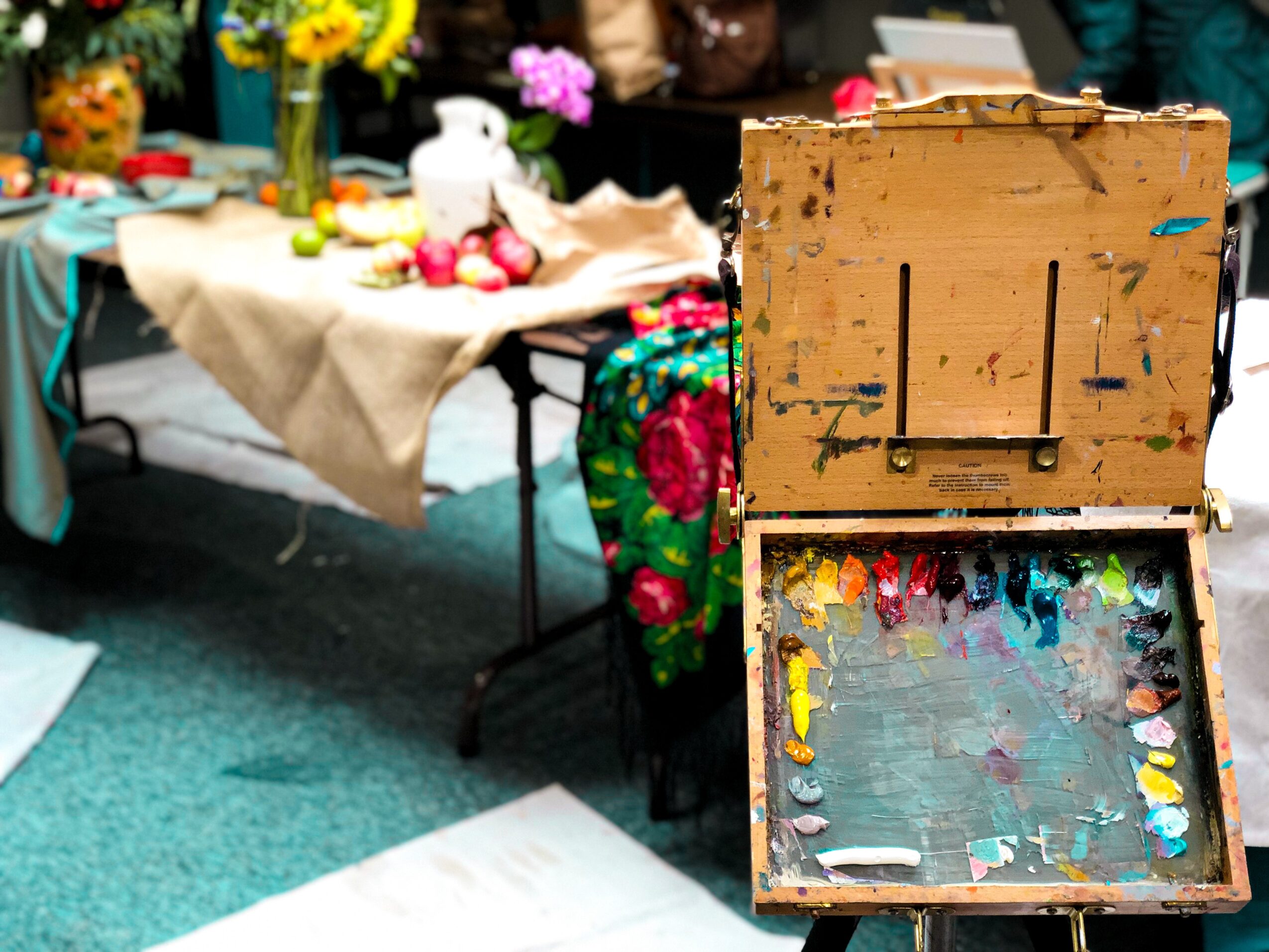 Open artist's palette with colorful paint, background shows a table with flowers and decorative items, at an indoor event.