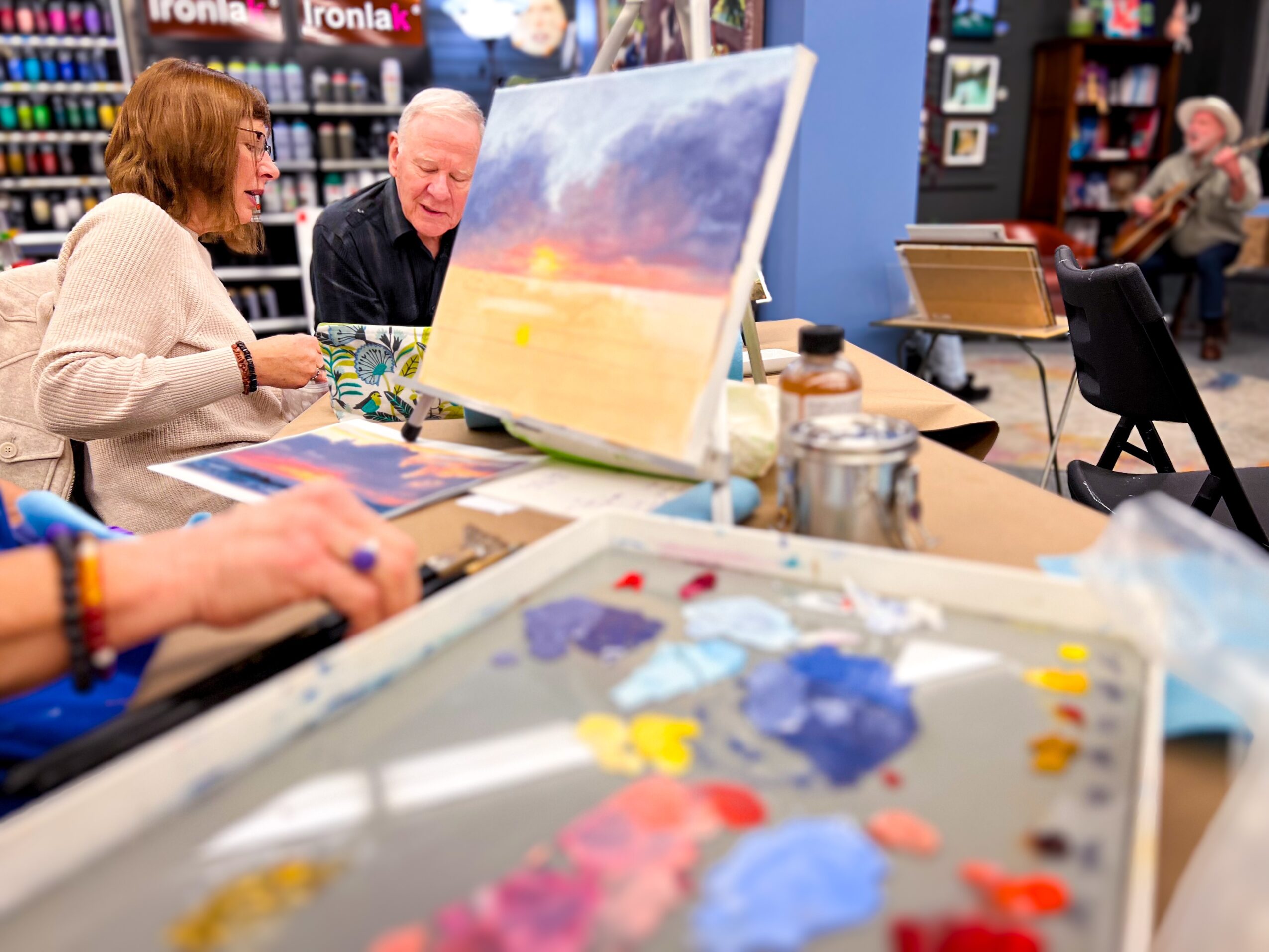 People painting and discussing artwork in a room with bookshelves, with a large canvas and art supplies visible.