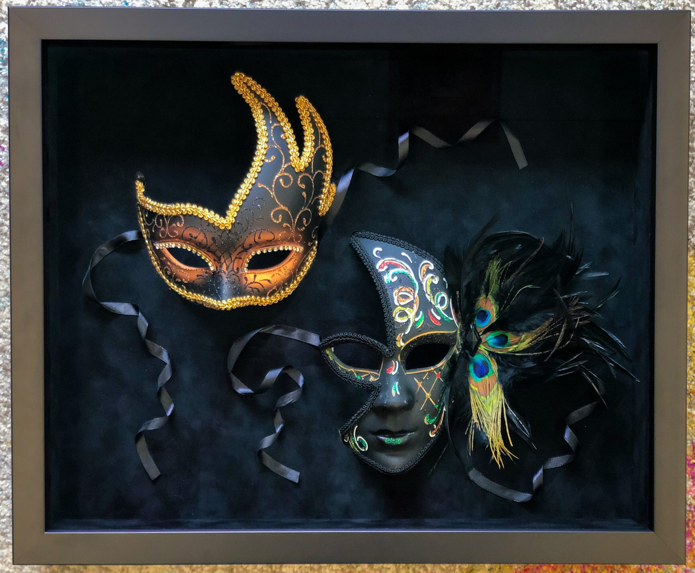 Two decorative masks with feathers and intricate designs, displayed against a dark background, with ribbons around them.