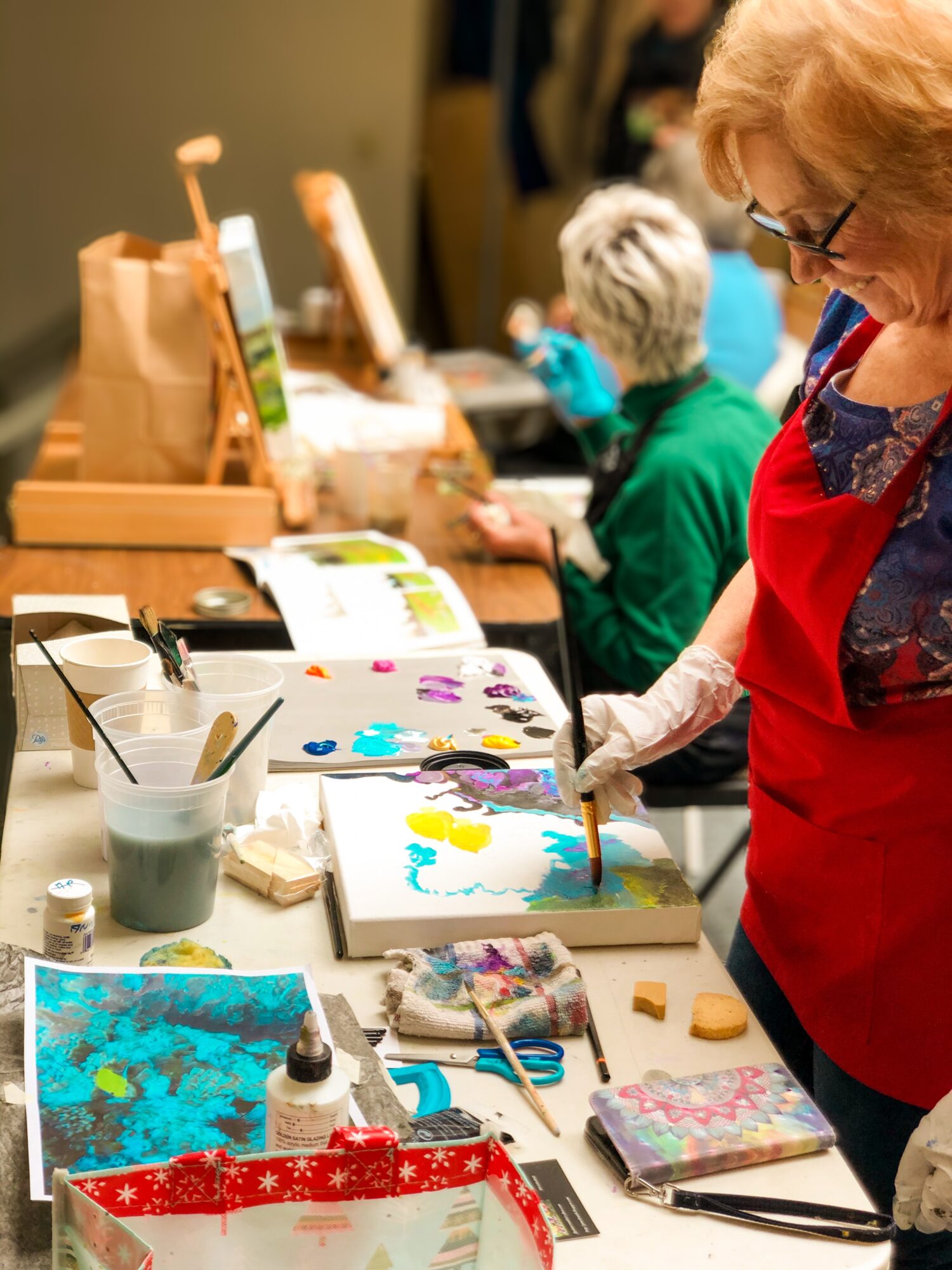 People painting on canvases at a table with art supplies, including brushes, paints, and paper, in an indoor setting.