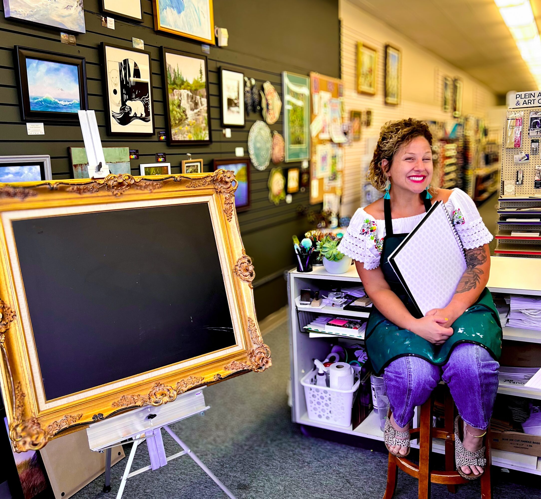 Smiling woman sitting on a stool in an art store, holding a clipboard, surrounded by artwork and supplies.