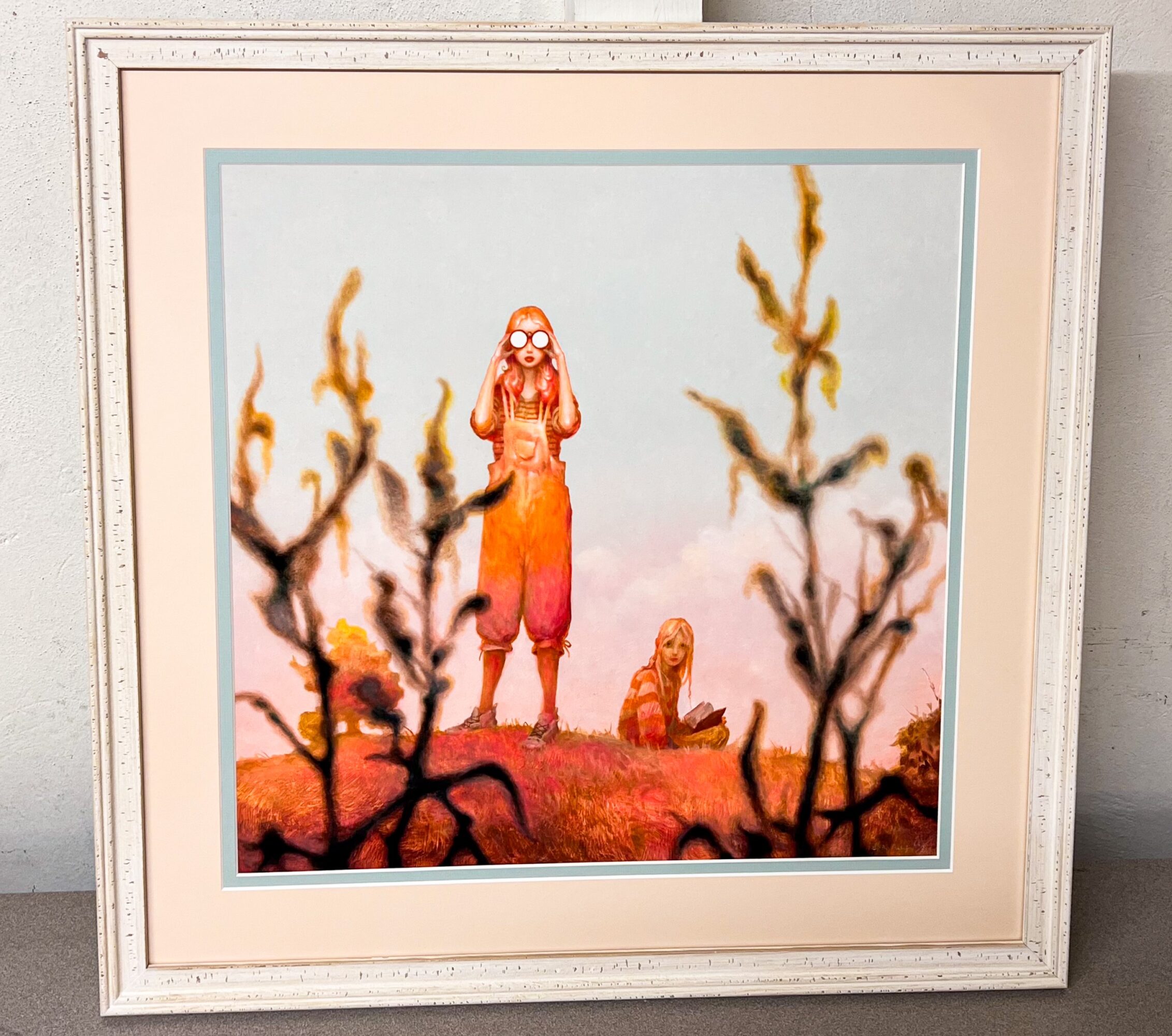 Framed artwork depicting two characters in a surreal landscape with twisted trees and pastel sky, one standing and one sitting.