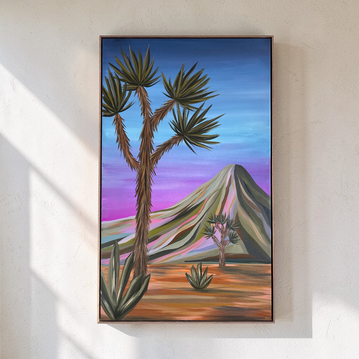 Painting of desert landscape with a large tree, mountain, and colorful sky, framed and hung on a wall.