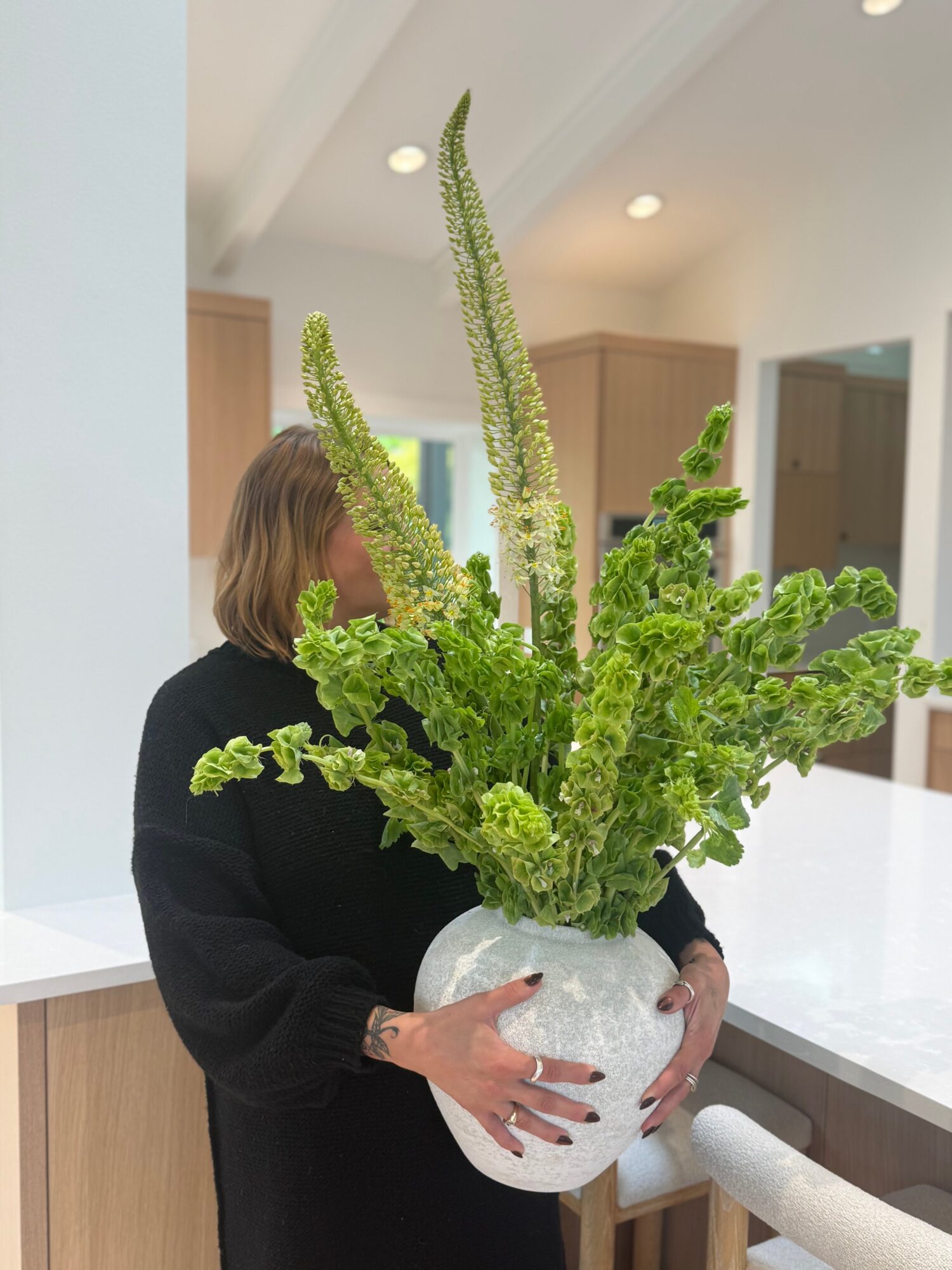 Person holding a large white vase with green, textured, elongated and curly plants inside, in a modern indoor space.