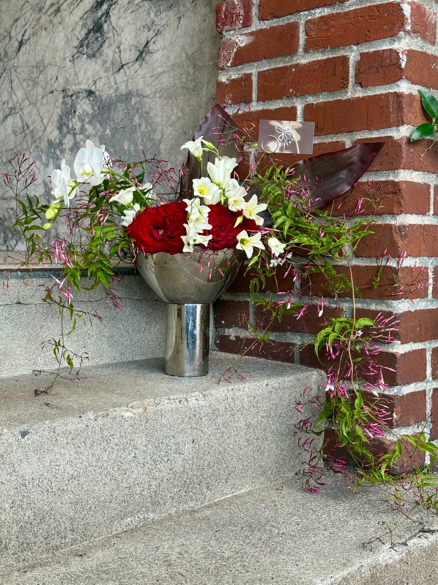 Flower arrangement with red and white flowers in a metal vase on concrete ledge against brick and stone wall