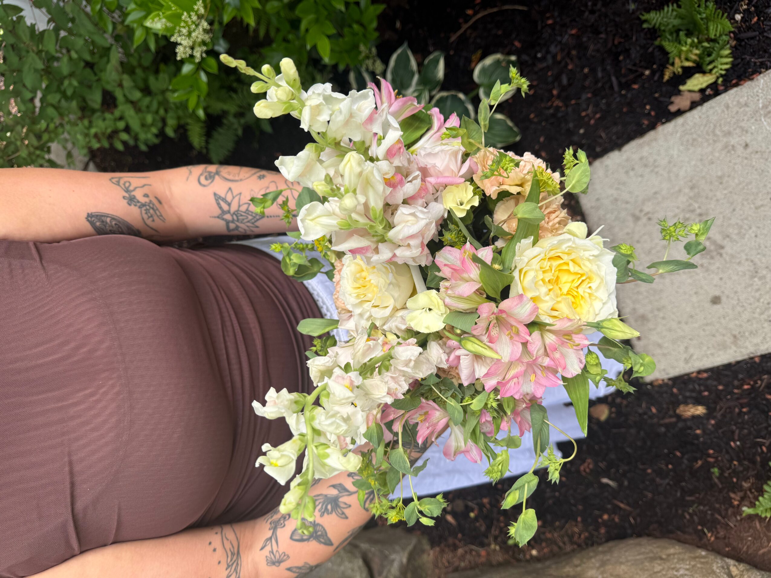 Person holding a large bouquet of white, pink, and yellow flowers outdoors, with greenery and a sidewalk in the background.