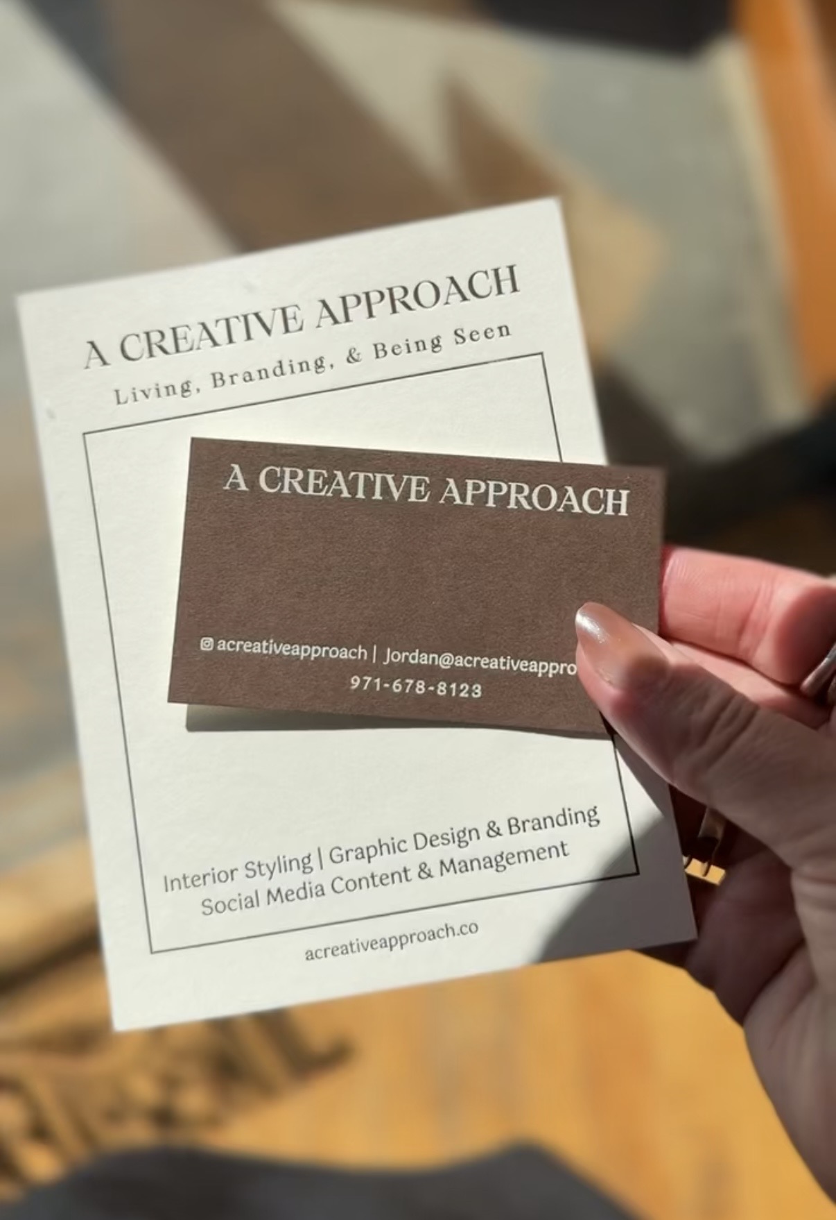 Person holding a dark business card with white text, with a white paper in the background on a wooden surface.