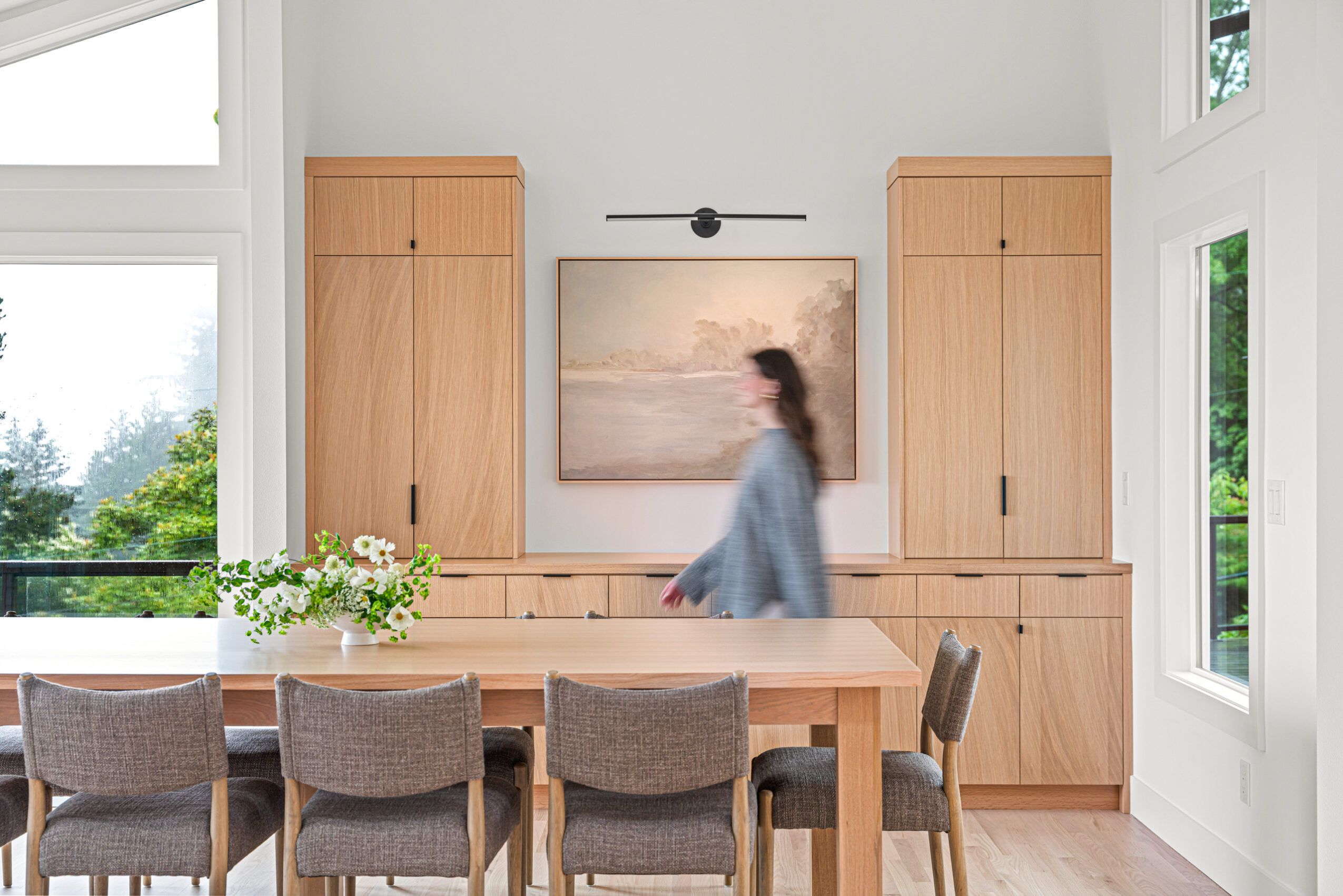 Person walking past a wooden dining room with large windows, a painting, and a table with a flower arrangement.