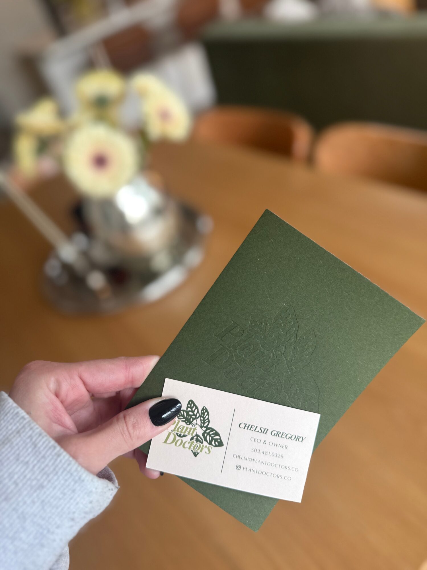 Person holding a green card and a white business card with a butterfly logo, in a room with a wooden table and flowers.