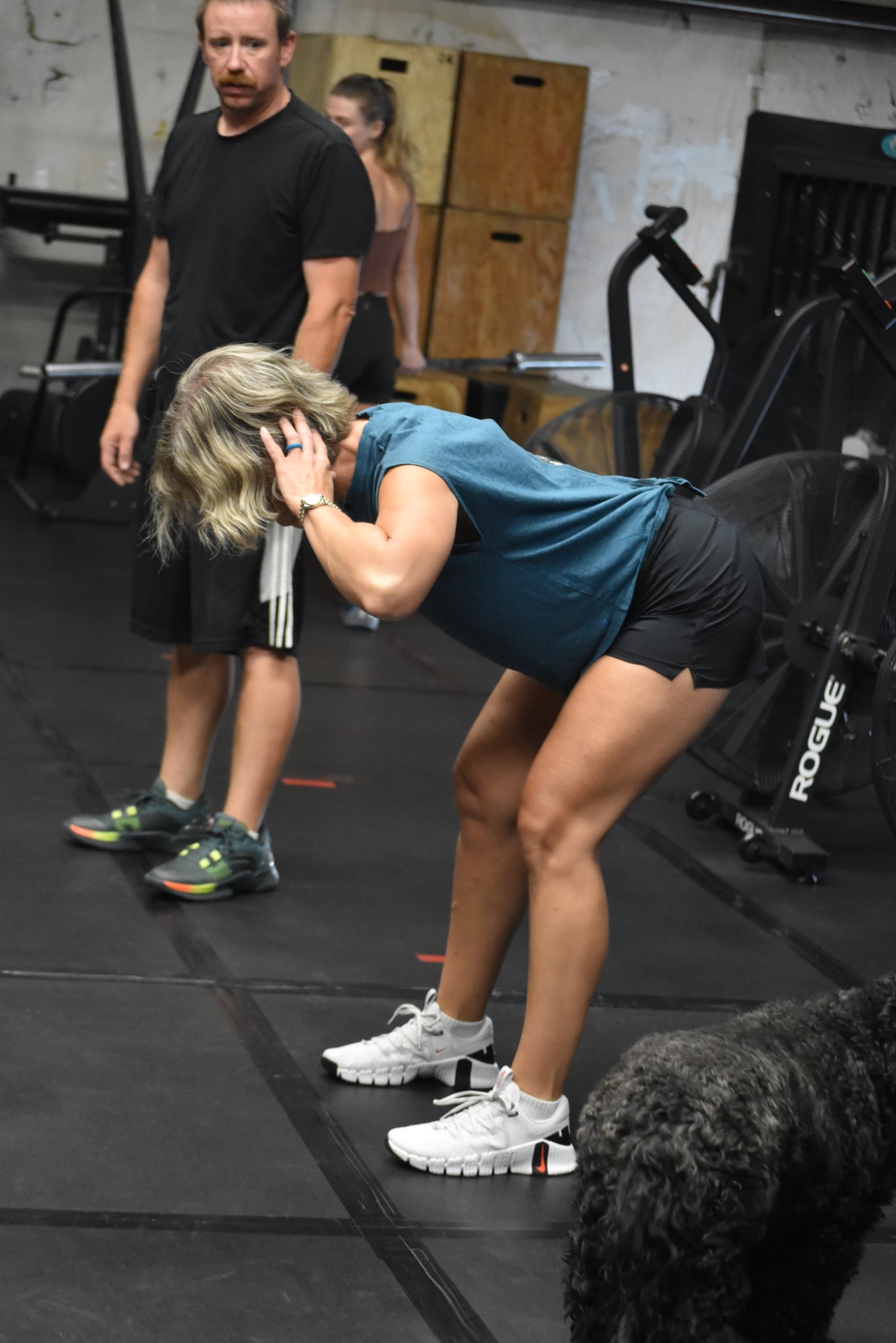 Woman in blue shirt and black shorts bends over, covering her ears, in a gym with two men nearby, one in black shirt.