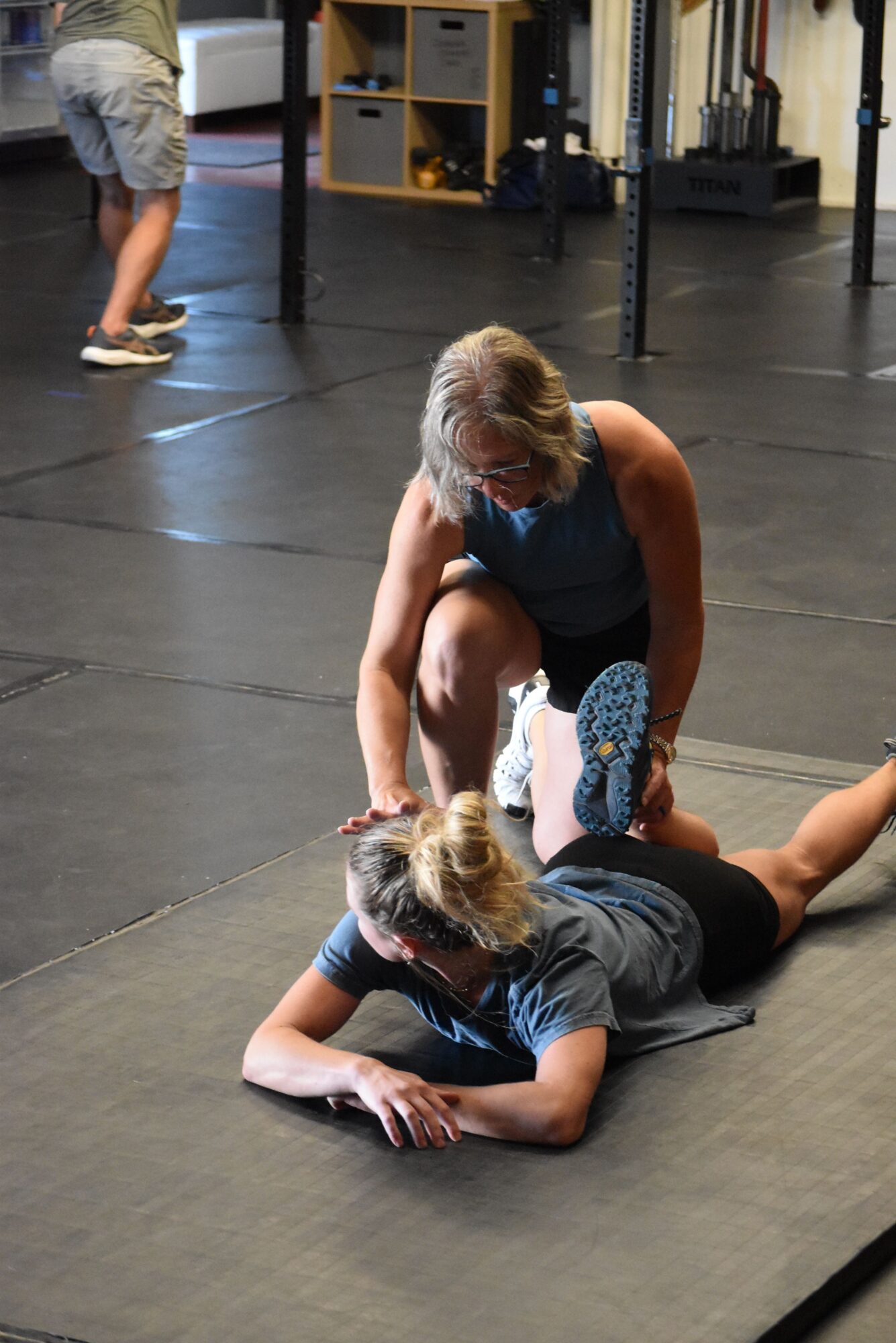 Two women practicing a fitness exercise on a gym floor, one standing and one lying on the ground, with gym equipment in the background.