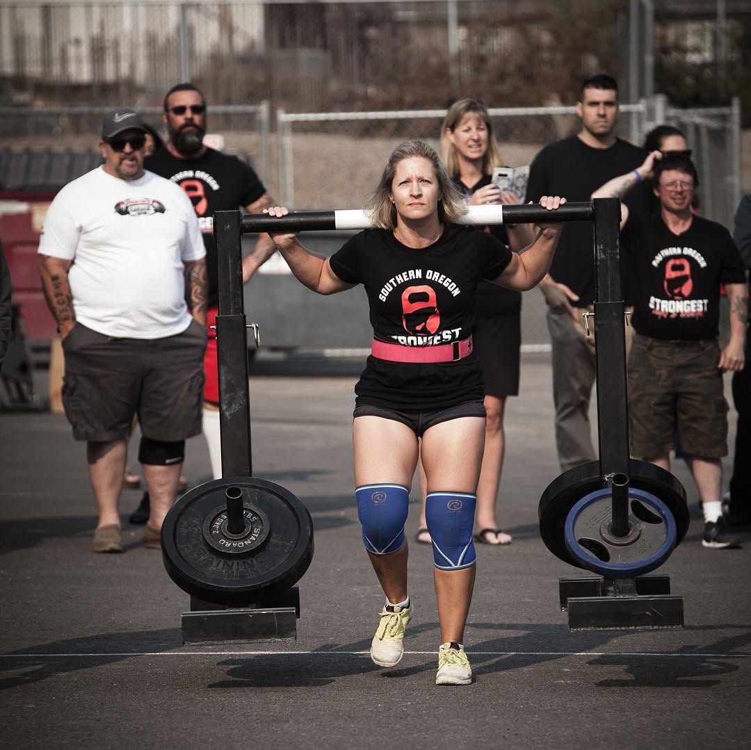 Woman lifting a barbell with weights at a gym, surrounded by people watching outdoors.