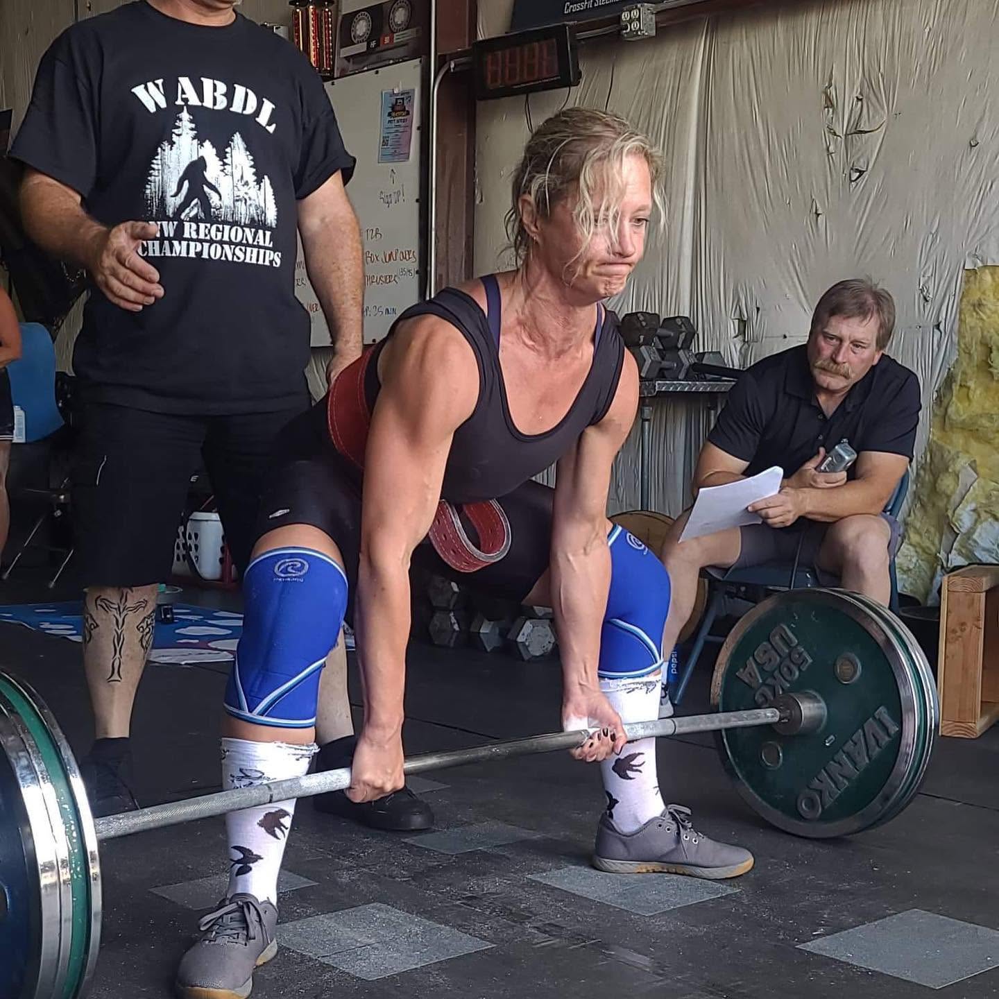 Woman lifting a barbell with weights, wearing blue knee sleeves and socks, in a gym setting. Two men are nearby, one holding papers.