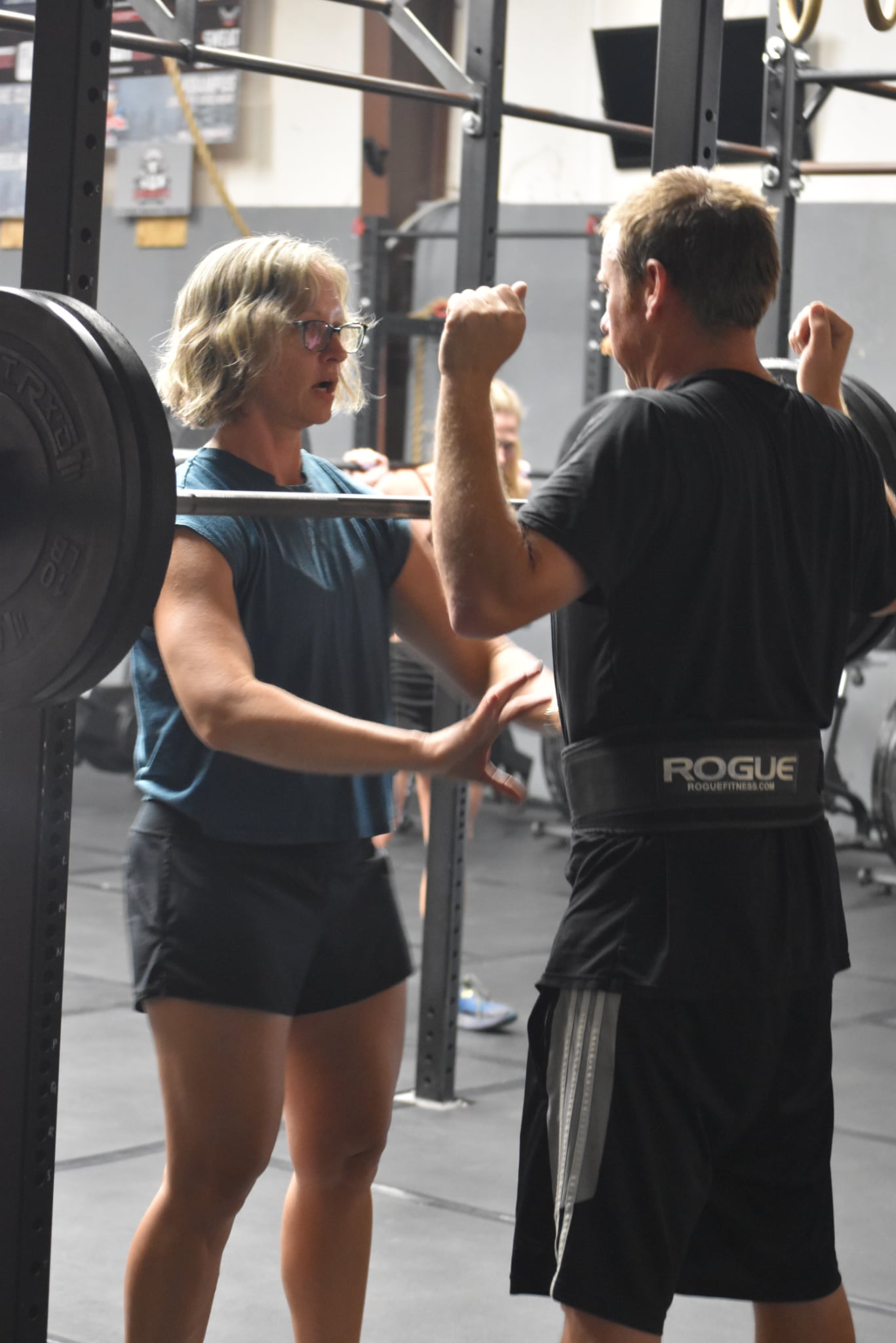 Two people in a gym, one woman and one man, engaging in a conversation or training, with gym equipment in the background.