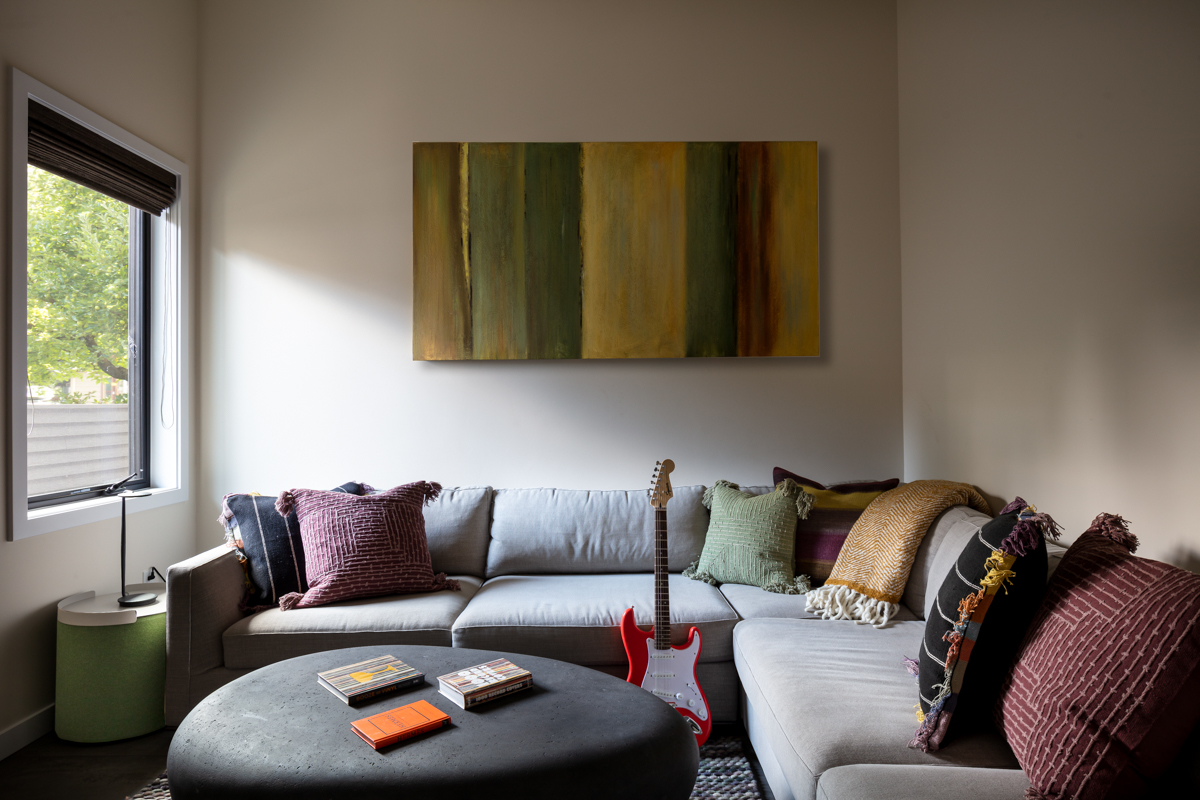 Living room with a large gray sectional sofa, colorful pillows, a black round coffee table, and a red guitar, with a window on the left and an abstract painting above the sofa.