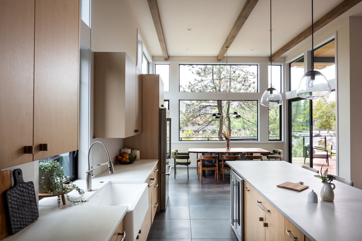 Modern kitchen with white countertops, wooden cabinets, and large windows overlooking trees and outdoor seating area.