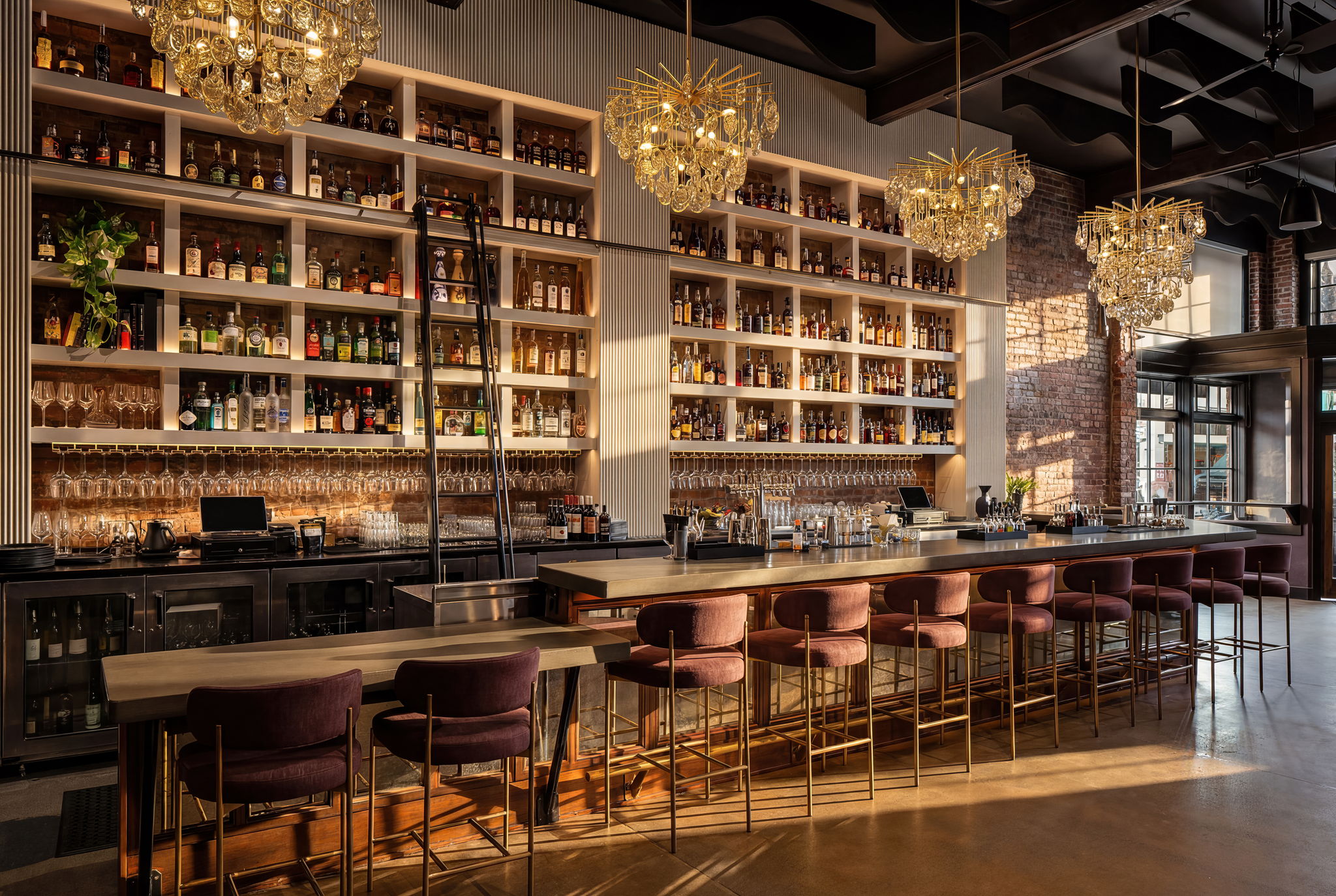 Bar with shelves of bottles, chandeliers, and a long counter with chairs, illuminated by natural light.
