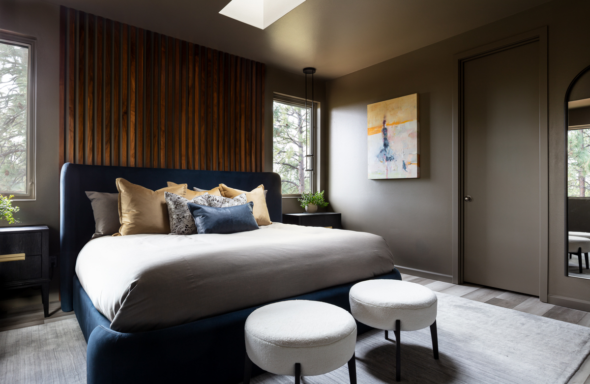 Bedroom with a large bed, dark headboard, and multiple pillows, two white ottomans in front, window with curtains, and artwork on wall.