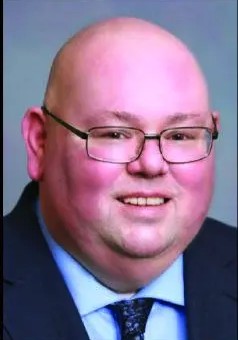A person with glasses, smiling, wearing a suit and tie, with a plain background.