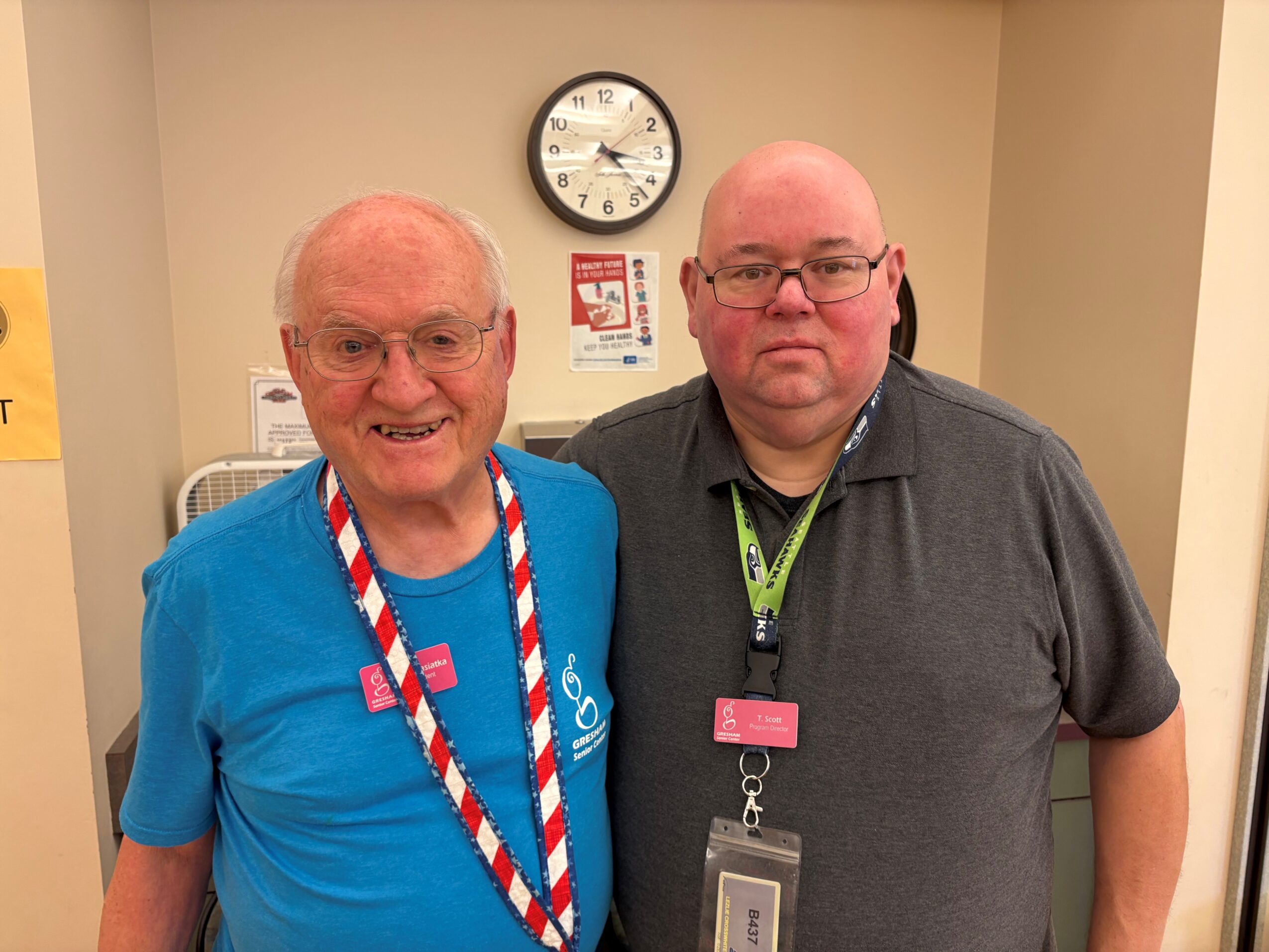 Two men standing indoors in front of a beige wall with a clock, smiling, one wearing a blue shirt and the other a gray shirt.