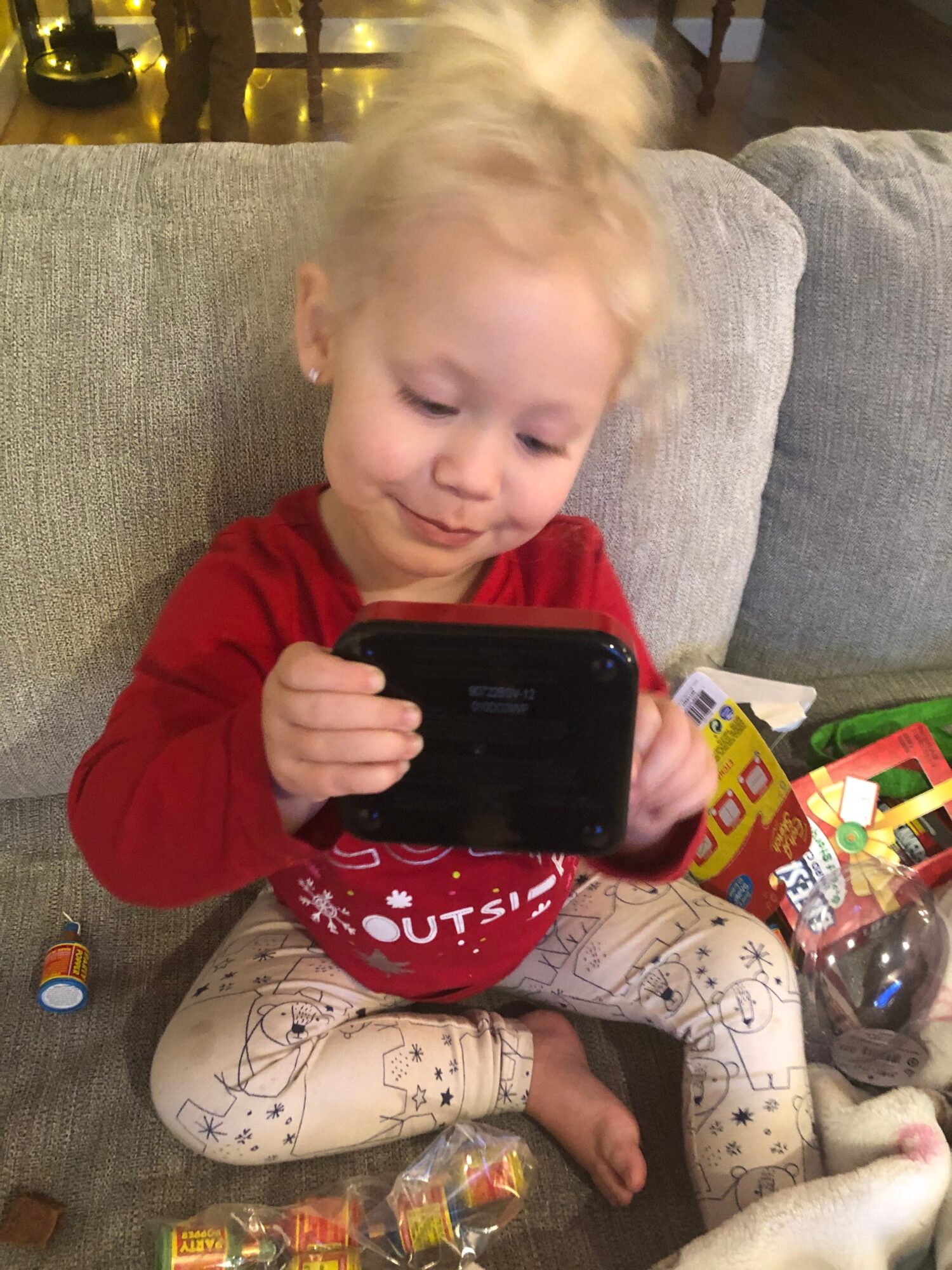 Child with blonde hair lying on a couch, holding a phone, surrounded by toys and snacks.