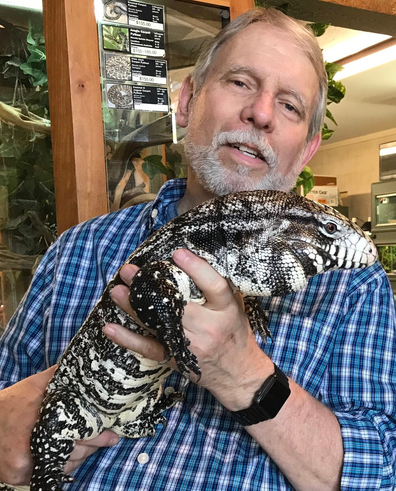 Man holding a large black and white patterned lizard indoors, smiling, wearing a blue checkered shirt and a black watch.
