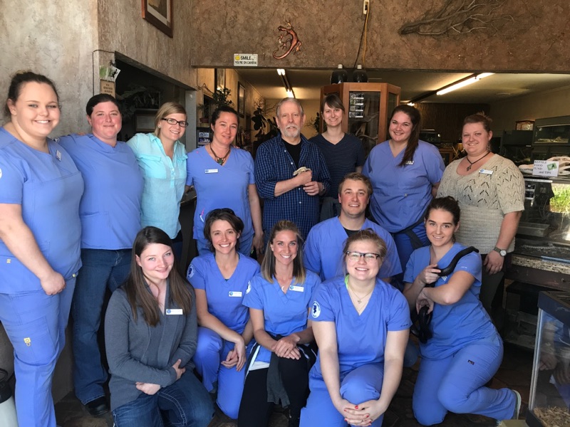 Group of healthcare workers in blue scrubs and uniforms standing together indoors, smiling, in a medical or hospital setting.