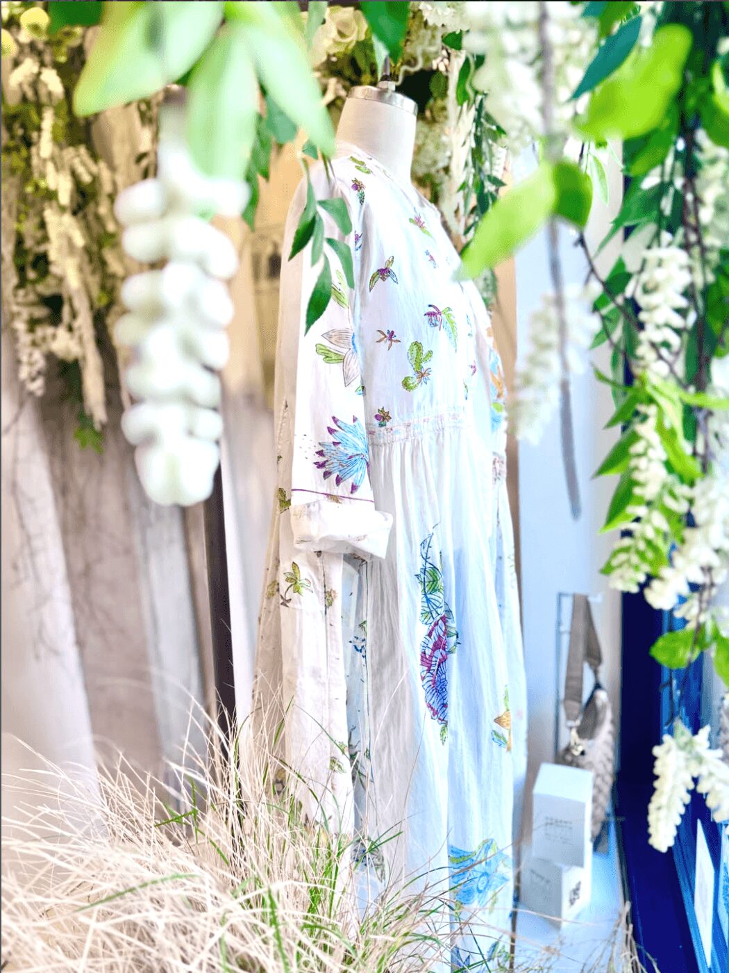 White dress with floral patterns displayed on a mannequin among green leaves and white flowers.