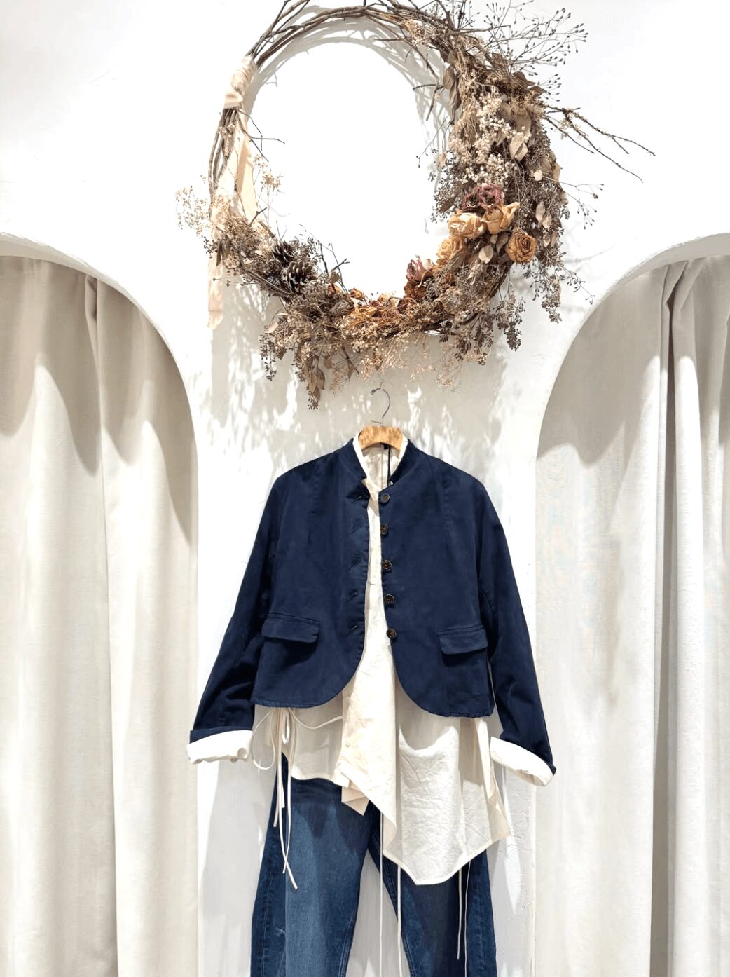 Clothing display with a navy jacket over a white shirt, hanging beneath a decorative wreath of dried flowers and branches.