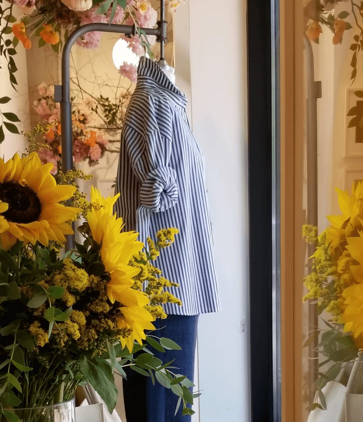 Side view of a striped shirt on a mannequin, surrounded by sunflowers and floral arrangements, near a window.