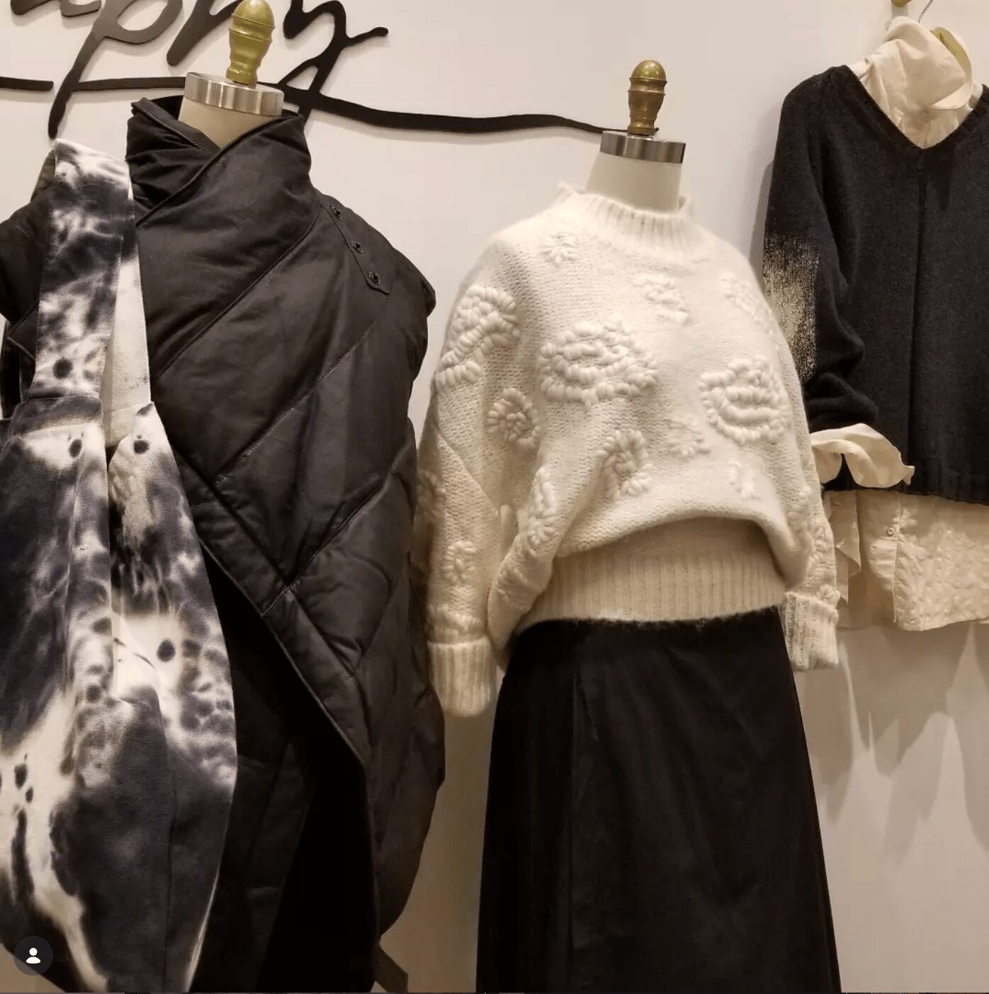Display of mannequins dressed in winter clothing, including a white textured sweater and a black skirt, with other garments nearby.