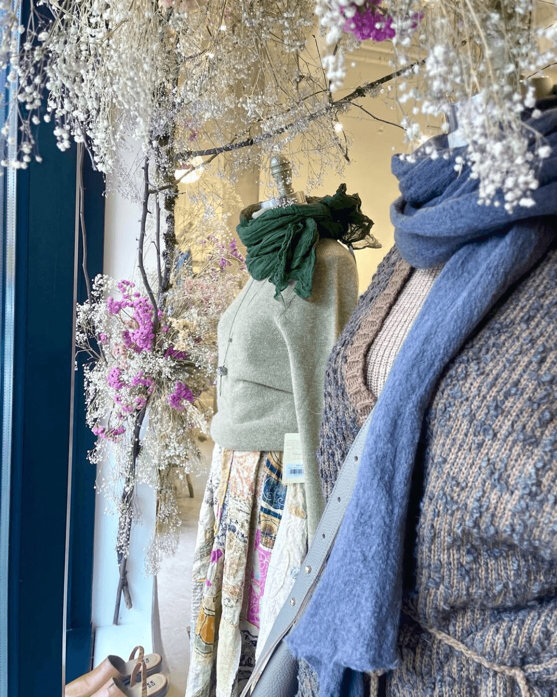 Three mannequins dressed in sweaters and scarves are displayed among floral arrangements with white and purple flowers.