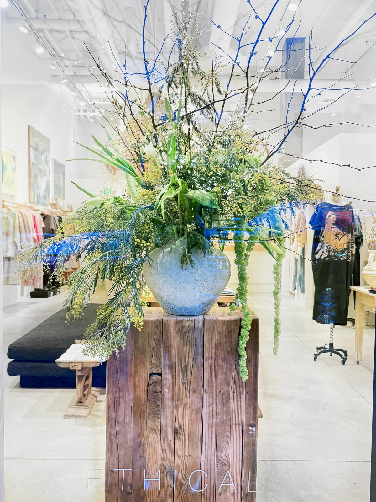 Large floral arrangement in a blue vase on a wooden pedestal inside a store, with clothing and furniture in background.
