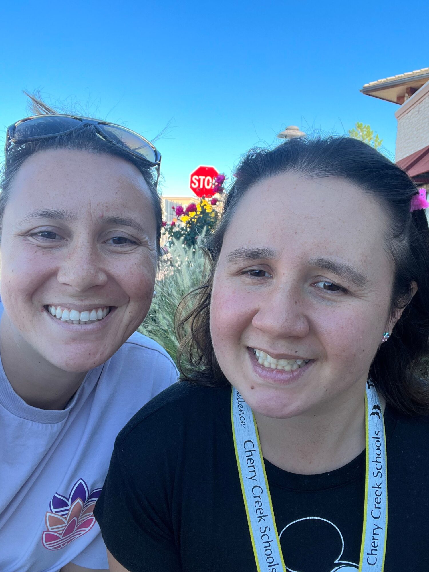 Two smiling women outdoors with blue sky, trees, and a sign in the background.