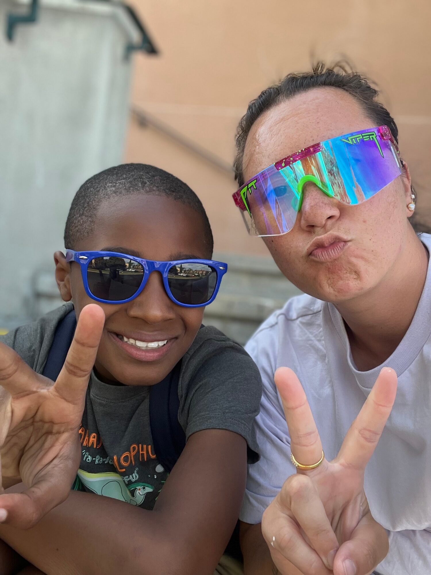 Two people wearing sunglasses making peace signs, smiling, outdoors, with a building in the background.