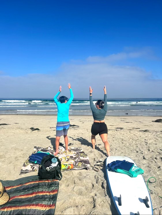 Two people stand on the beach facing the ocean with arms raised, wearing swimwear, under a blue sky with some clouds.