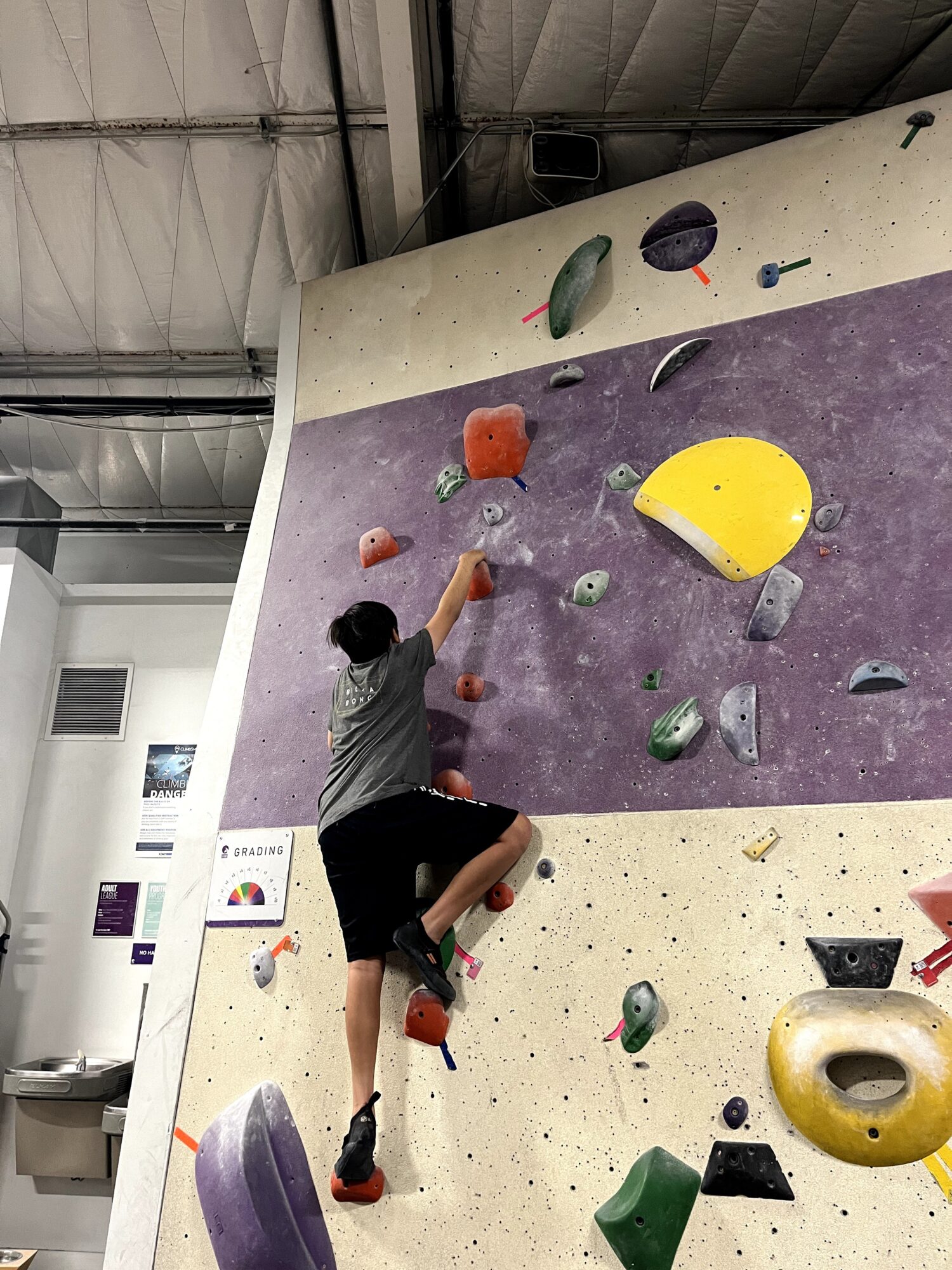 Child climbing on an indoor bouldering wall with colorful holds, reaching for a hold with one hand, in a gym.