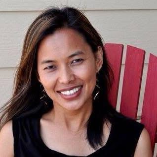 Woman with long dark hair smiling, sitting on a red wooden chair outdoors, wearing a black sleeveless top.