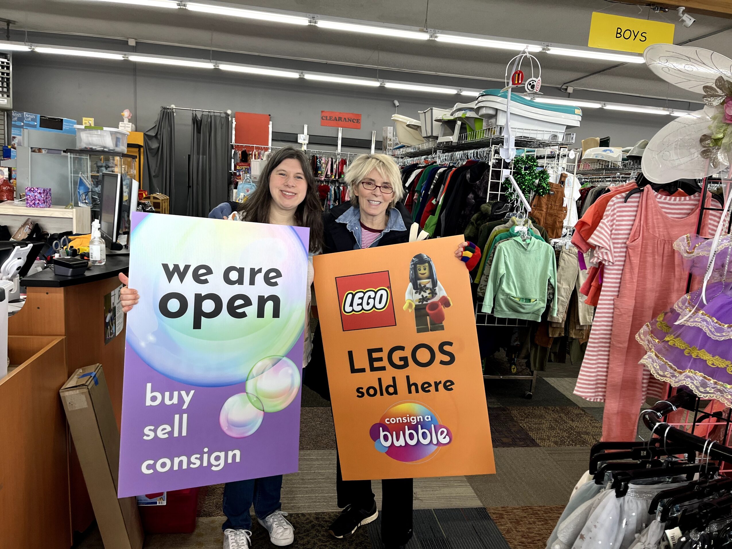 Two women holding signs inside a thrift store, surrounded by clothing and merchandise, smiling at the camera.