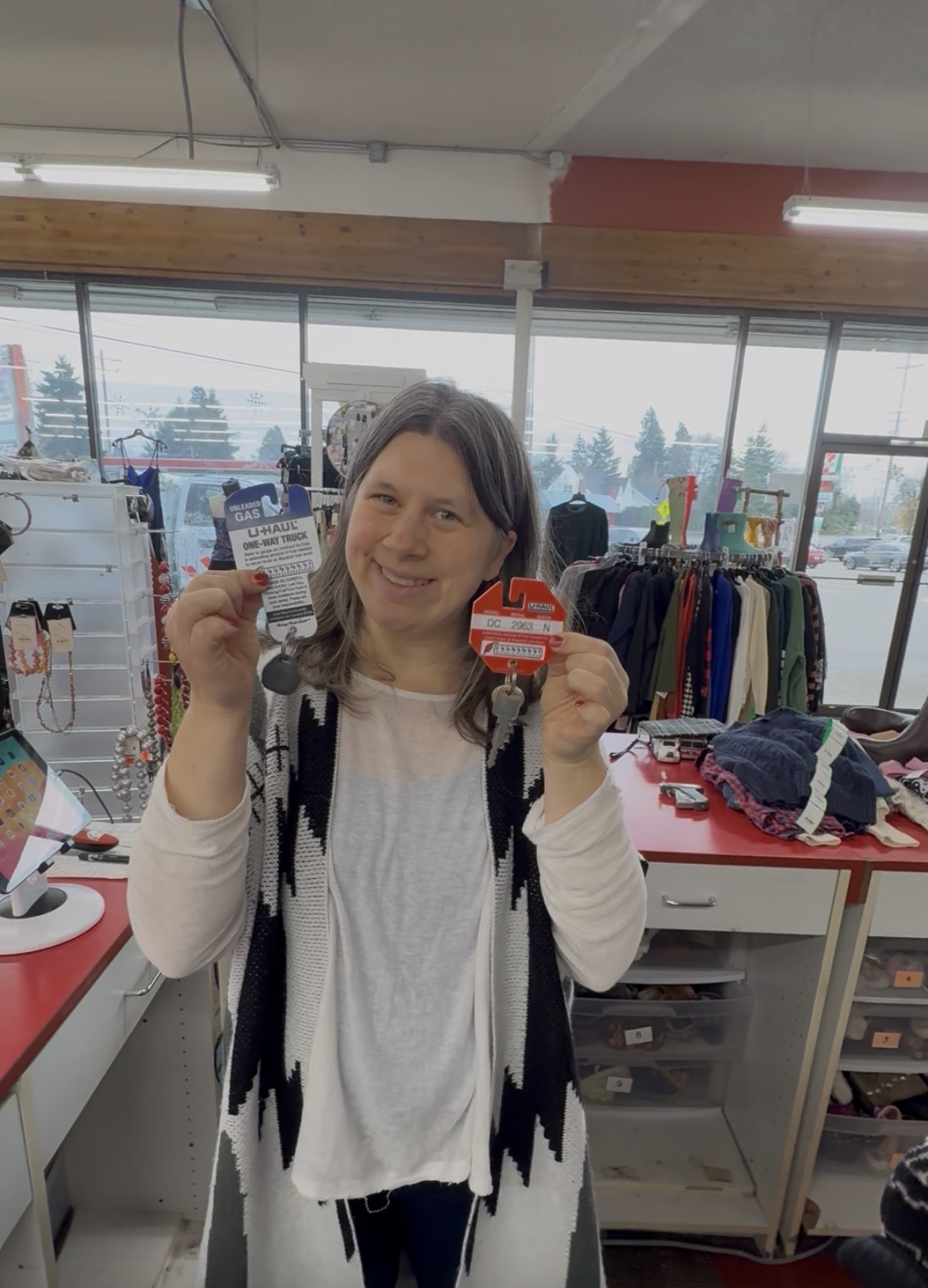 Woman holding a card and a red object inside a store, smiling, with clothing racks and windows in background.