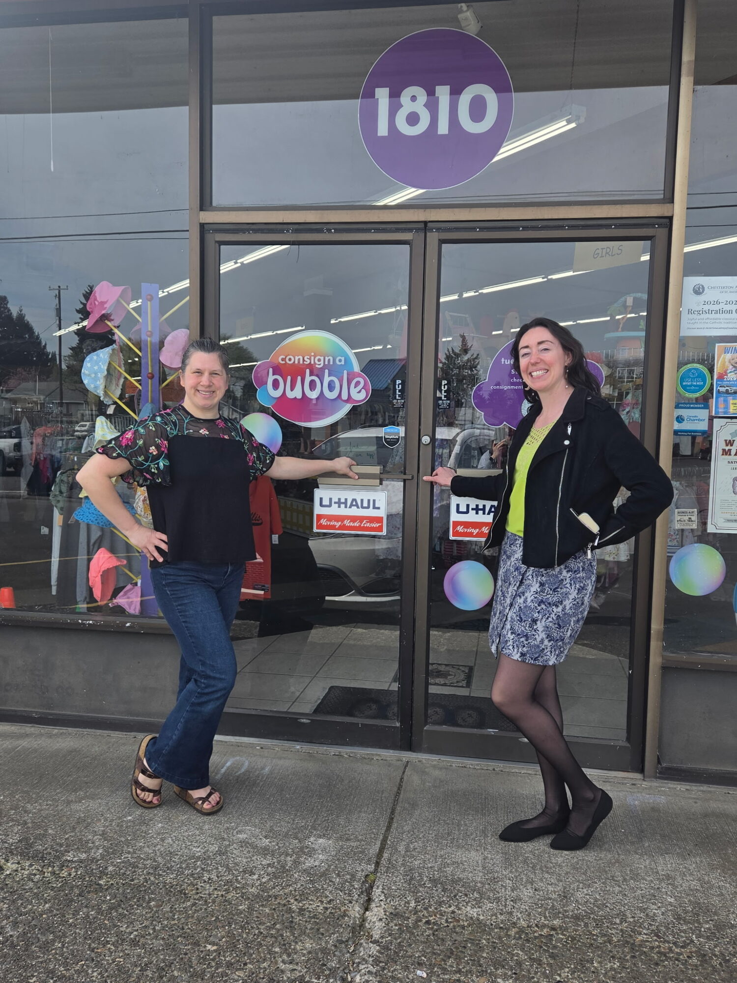 Two women stand outside a store with glass doors, smiling, one with hands on hips, the other with hands in pockets.
