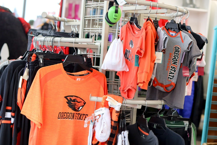 Clothing rack with Oregon State University sportswear, including orange shirts and gray backpacks, in a store or market setting.