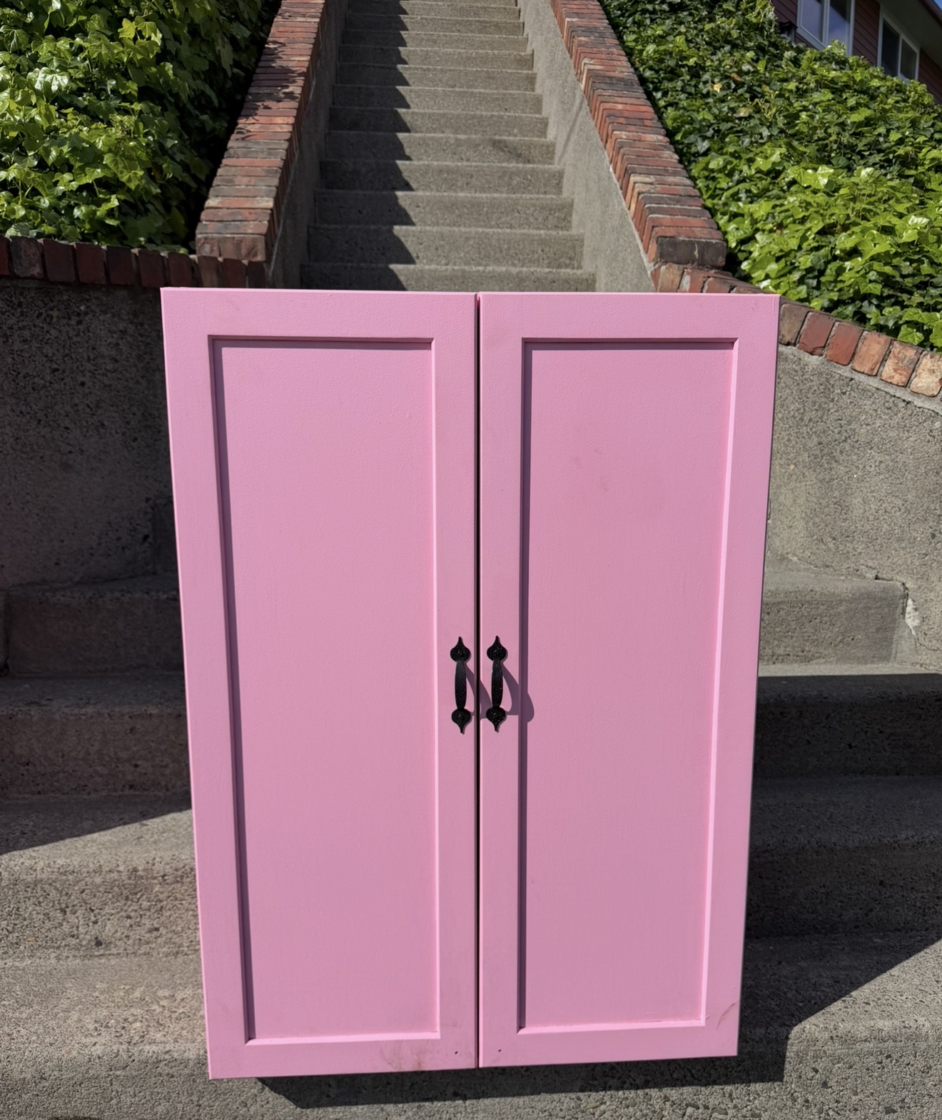 Pink cabinet with two doors and black handles, placed outdoors in front of stairs and greenery.