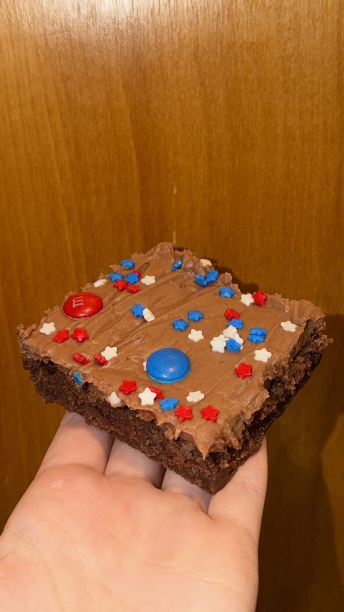 A hand holding a square chocolate brownie decorated with red, blue, and white sprinkles and candies, against a wooden background.