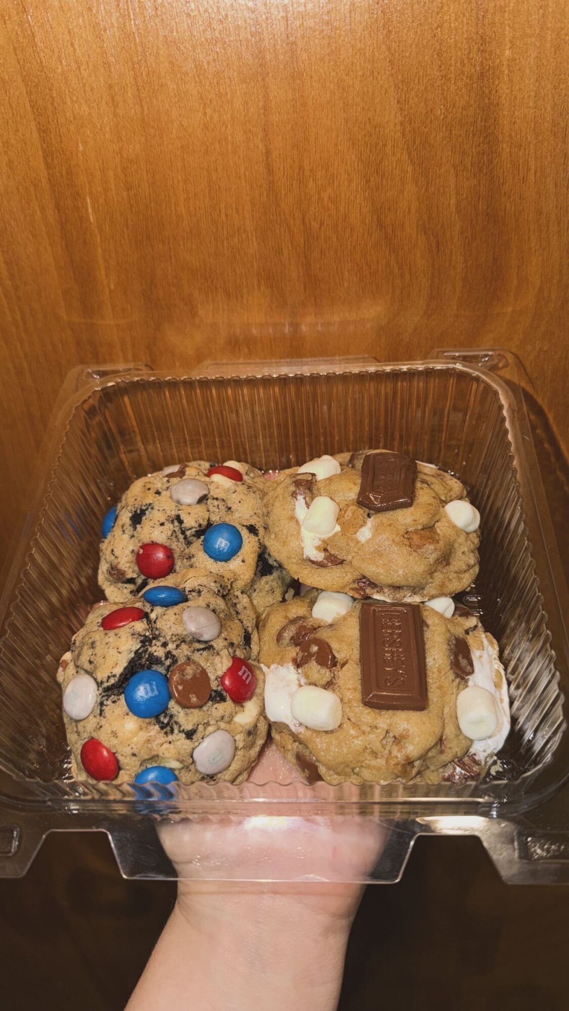 Four cookies with colorful candies and white chocolate chips in a clear plastic container, held by a hand against a wooden background.