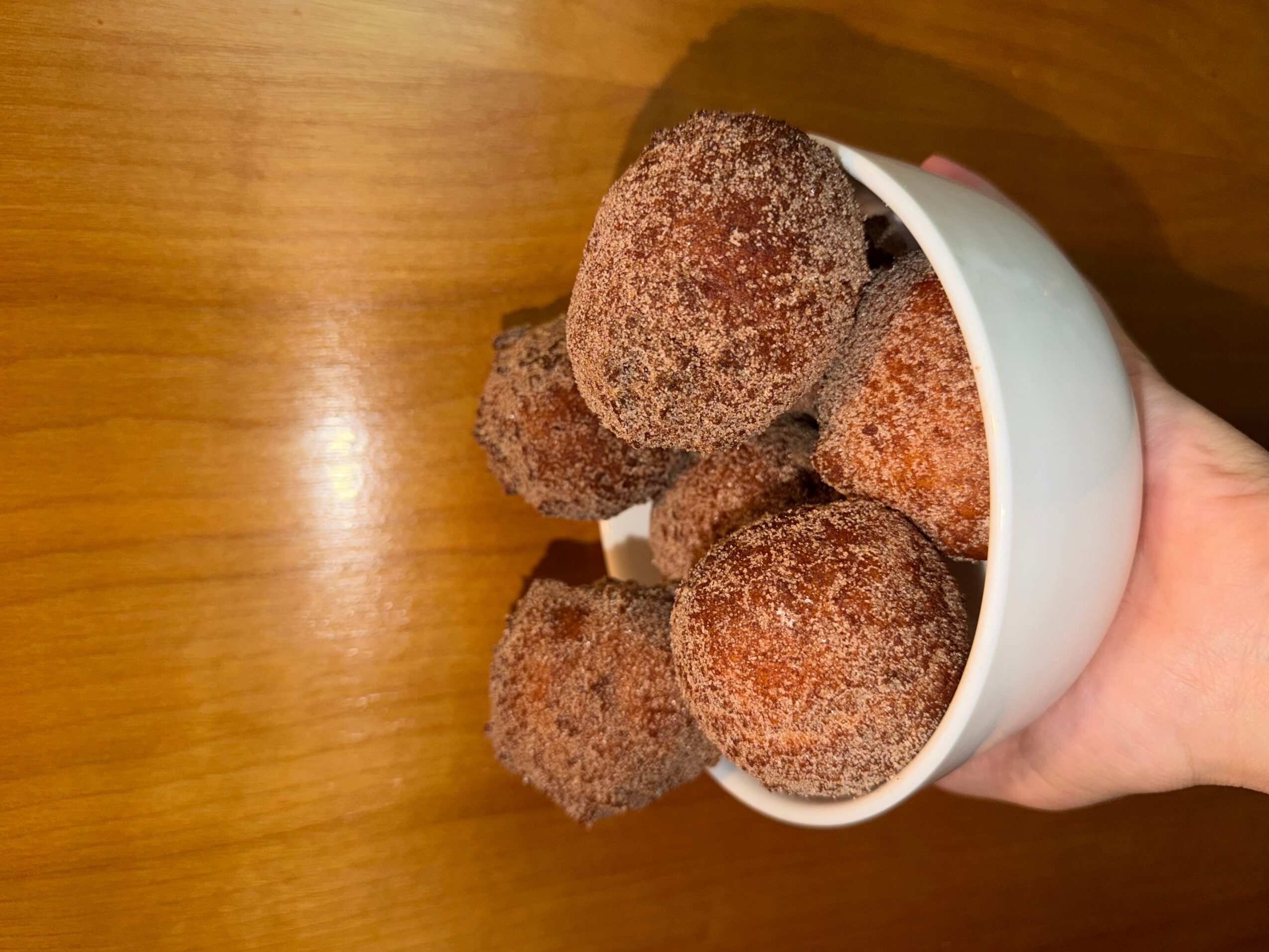 Hand holding a white cup filled with cinnamon-sugar coated donuts over a wooden surface.