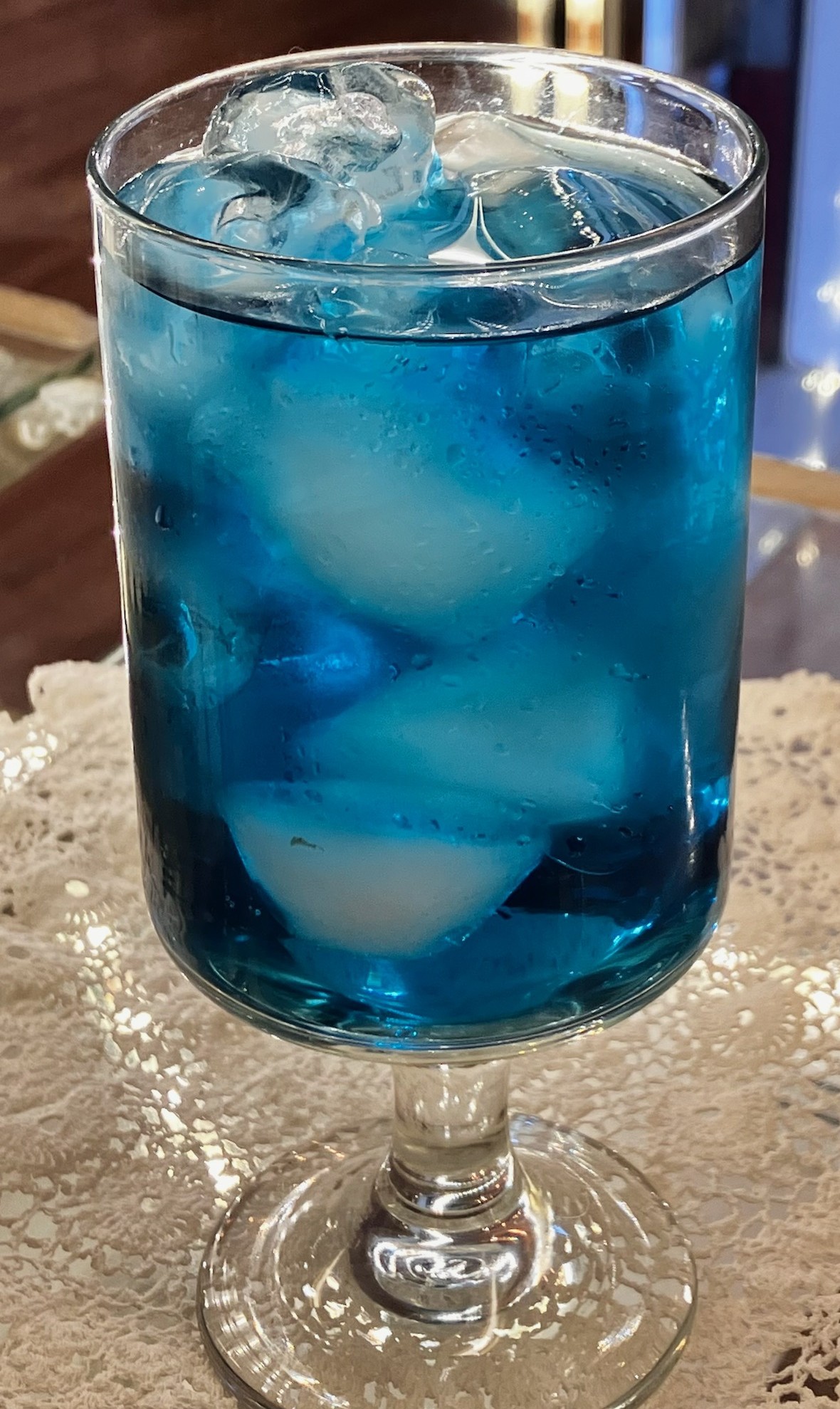 Tall glass filled with blue liquid and ice cubes on a lace tablecloth.