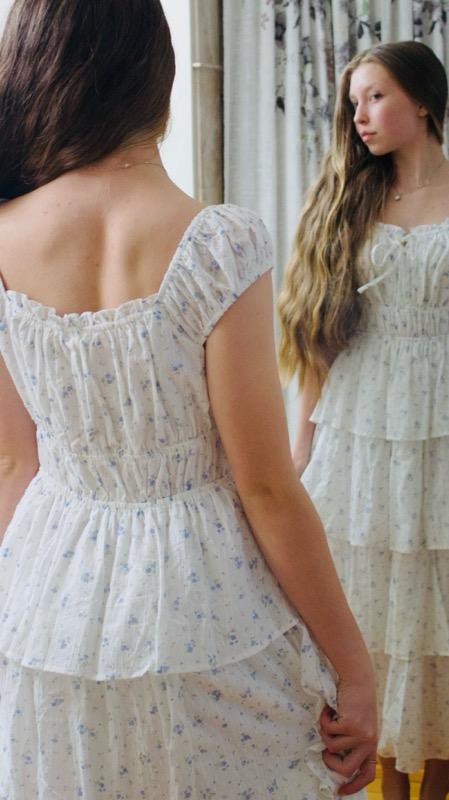 Young woman with long hair looking at her reflection in a mirror, wearing a white dress with a floral pattern.