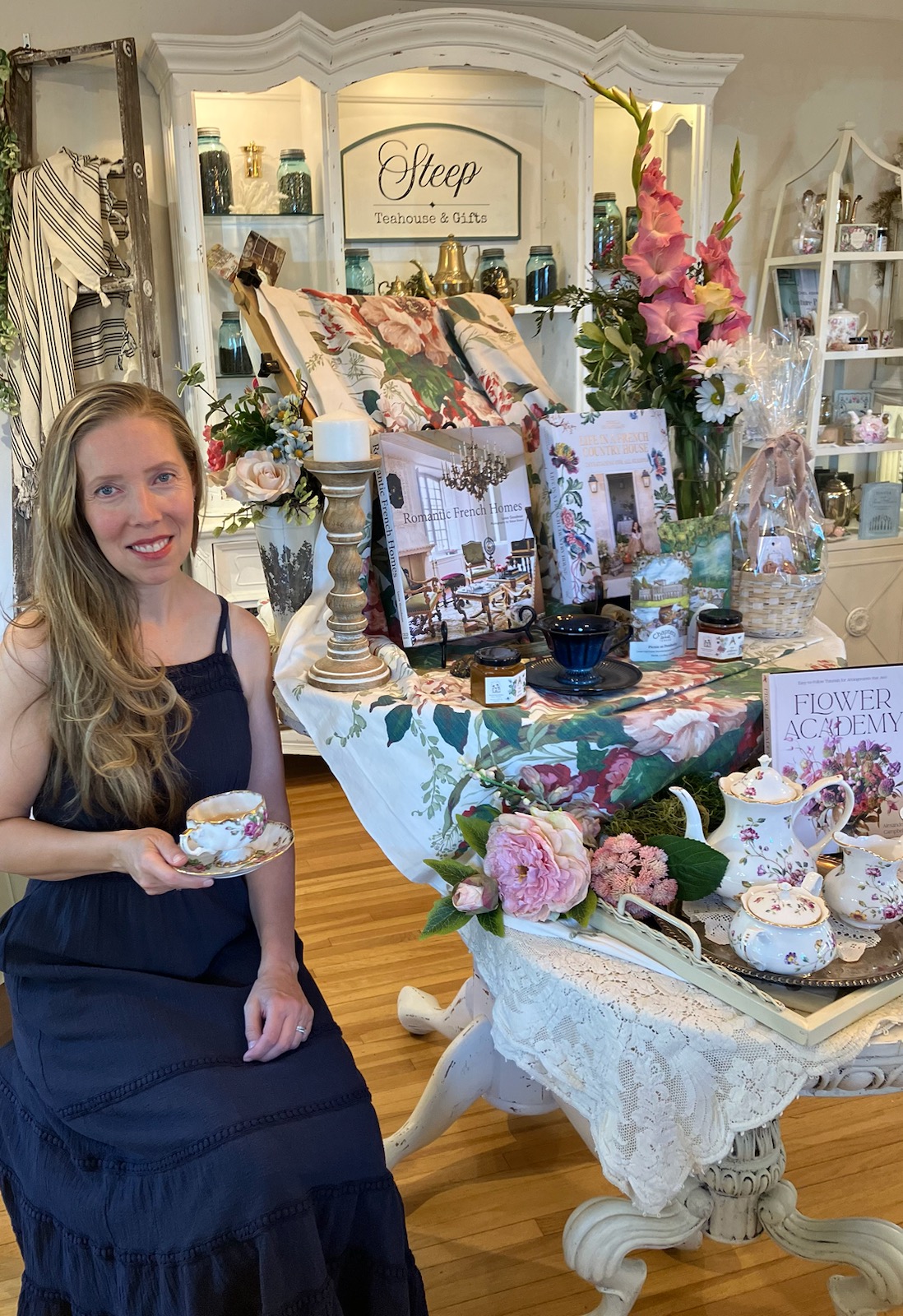 Woman with long hair in a dark dress holding a teacup, surrounded by floral arrangements and decorative items in a boutique.