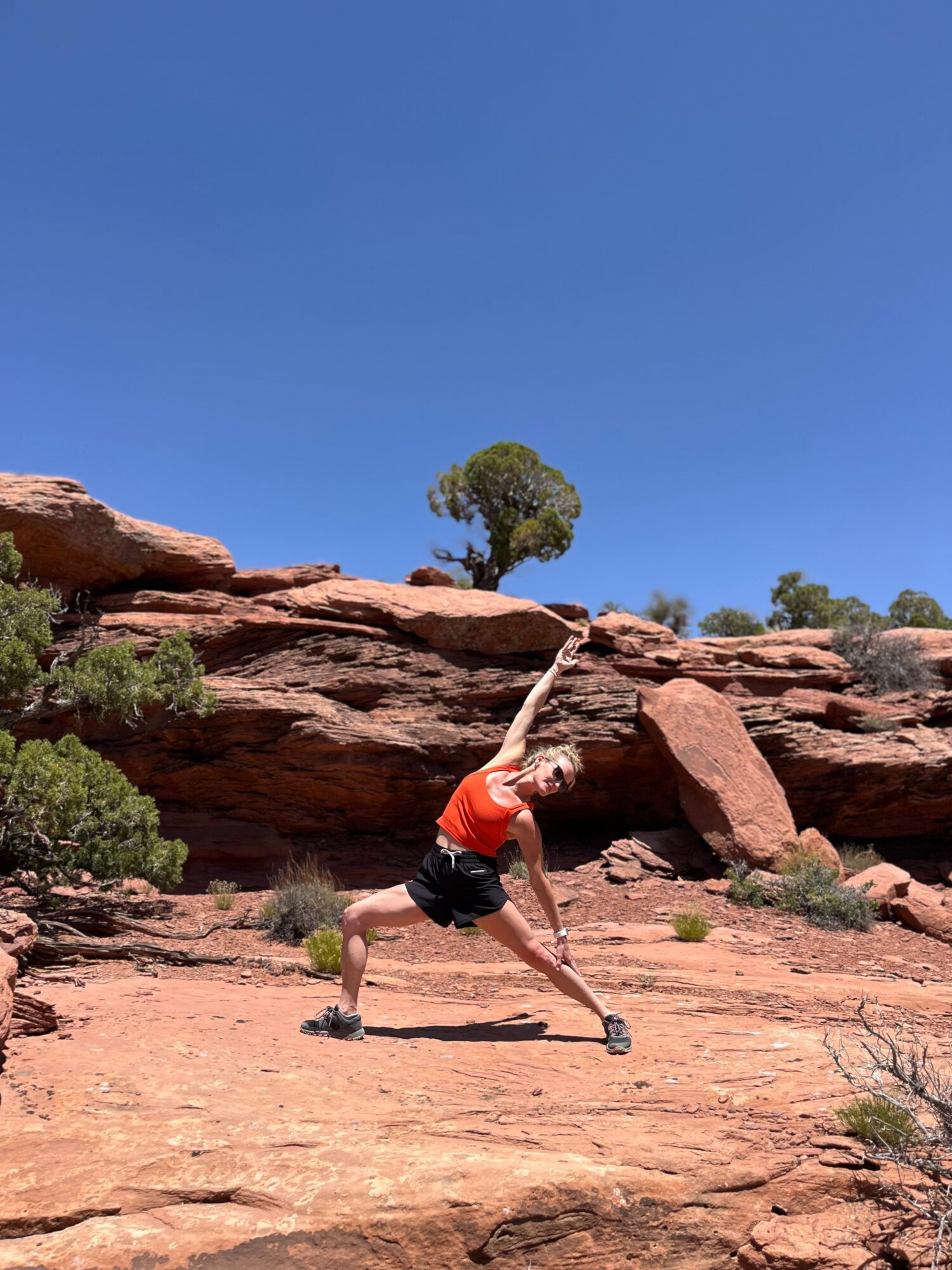 Person in orange shirt and black shorts stretching outdoors on rocky terrain with blue sky and trees in background.